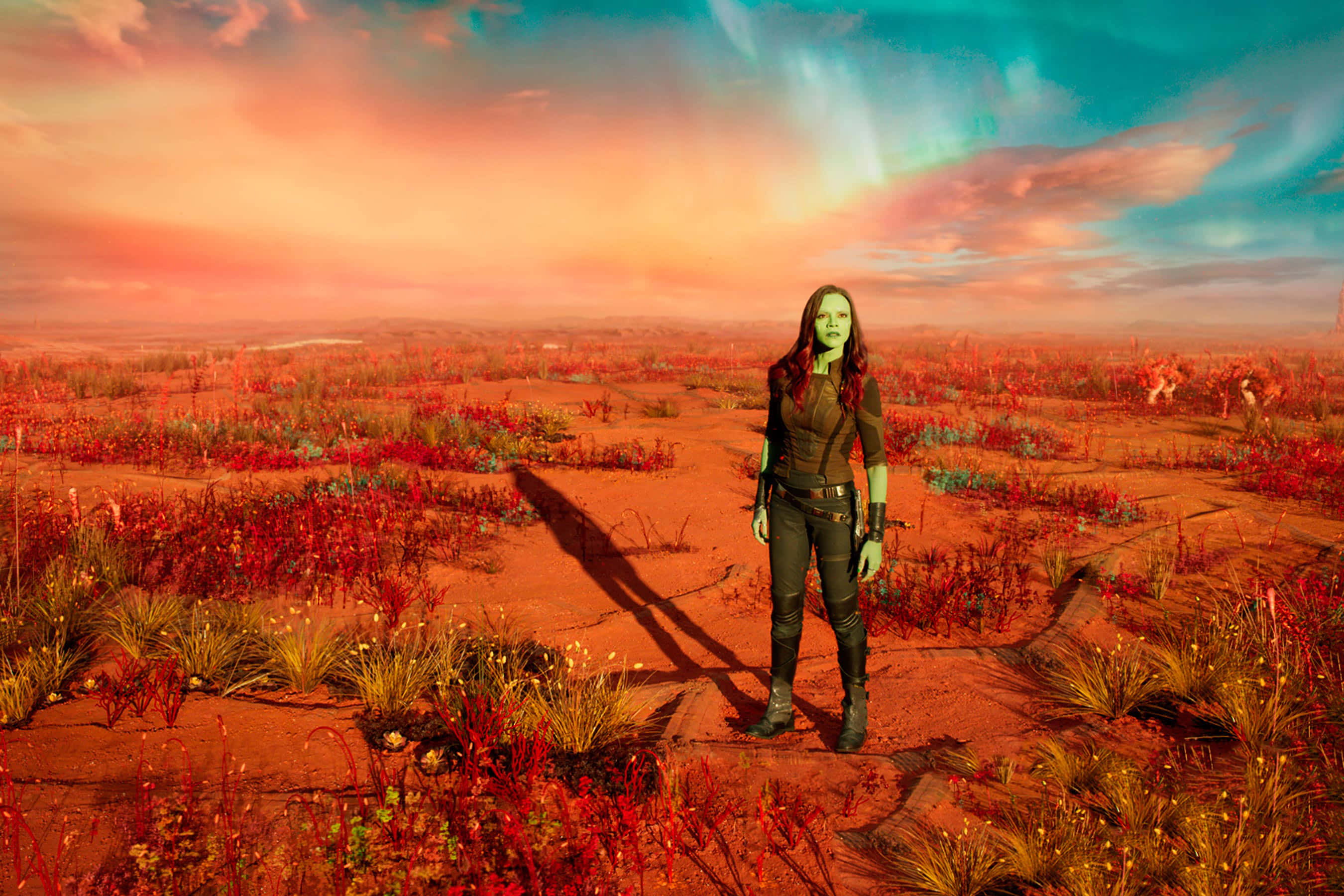 Guardians Of The Galaxy 2 桌布 壁紙 — 葛摩拉獨自站在超現實的外星沙漠中，天空是充滿活力的日落，有粉紅色和藍綠色的雲彩。