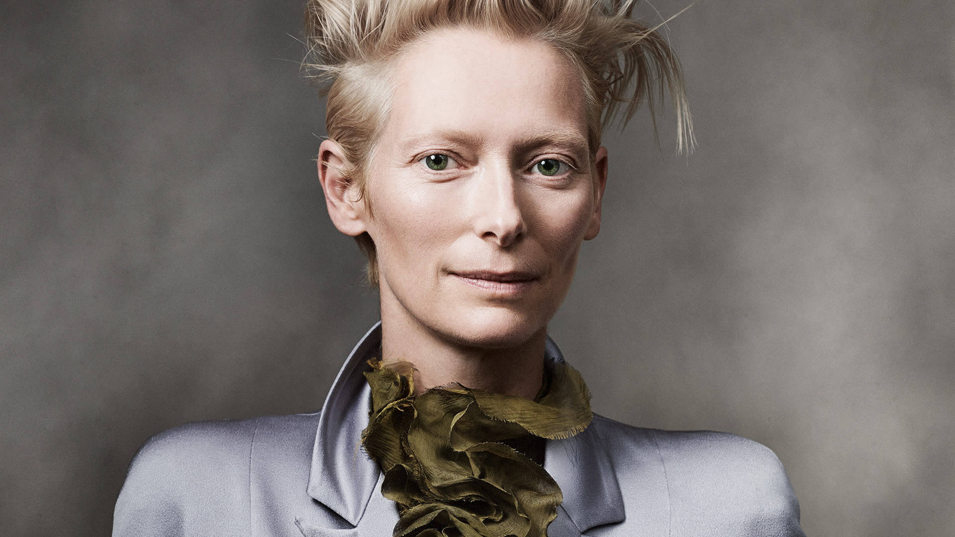 Celebrity 桌布 壁紙 — 蒂妲·史雲頓（Tilda Swinton）留著尖刺金髮和綠色眼睛，身穿淺灰色西裝，搭配橄欖色荷葉邊圍巾，背景為柔和的灰色。