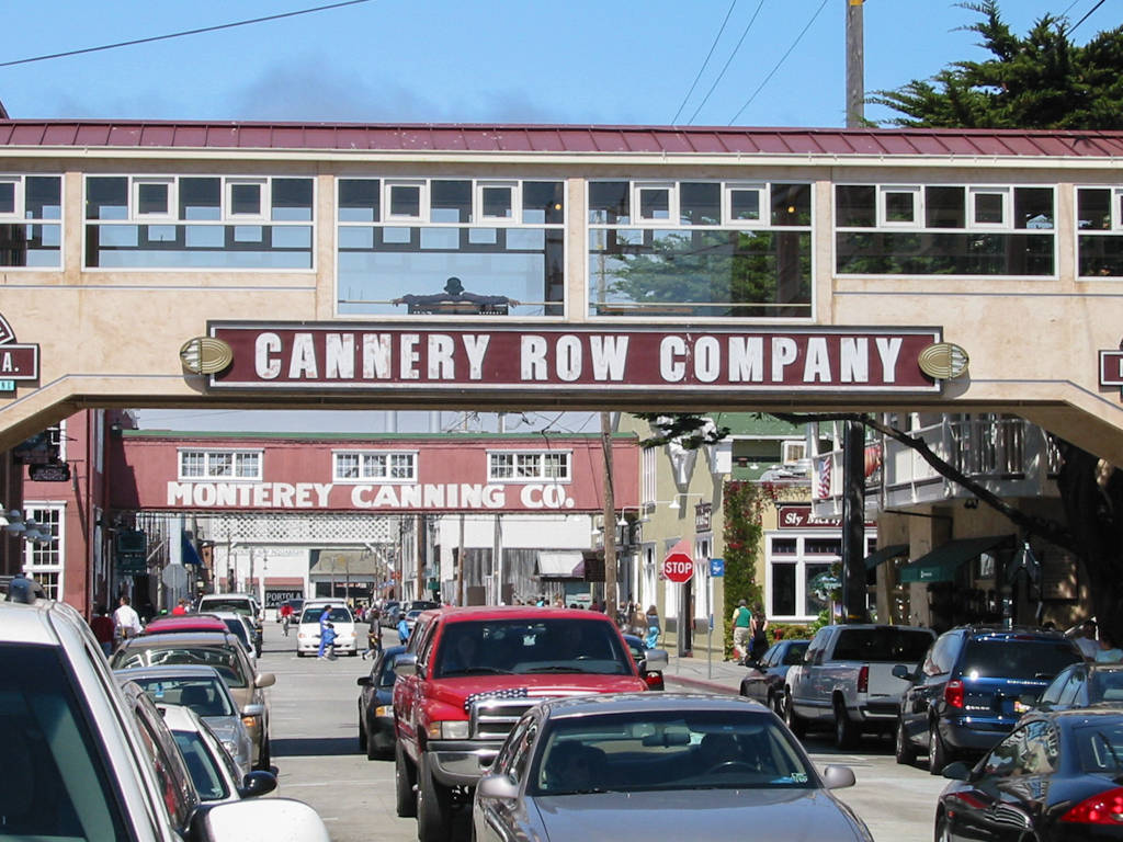 加州 桌布 壁紙 — 蒙特雷一條繁忙的街道，由一個帶有 'CANNERY ROW COMPANY' 標誌的大型拱門框住，天空清澈湛藍。