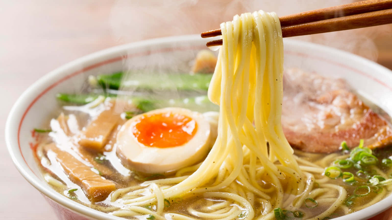 Noodles 桌布 壁紙 — 蒸氣從一碗熱騰騰的拉麵中升起，筷子夾起金黃色的麵條，湯上有半熟蛋和蔥花。