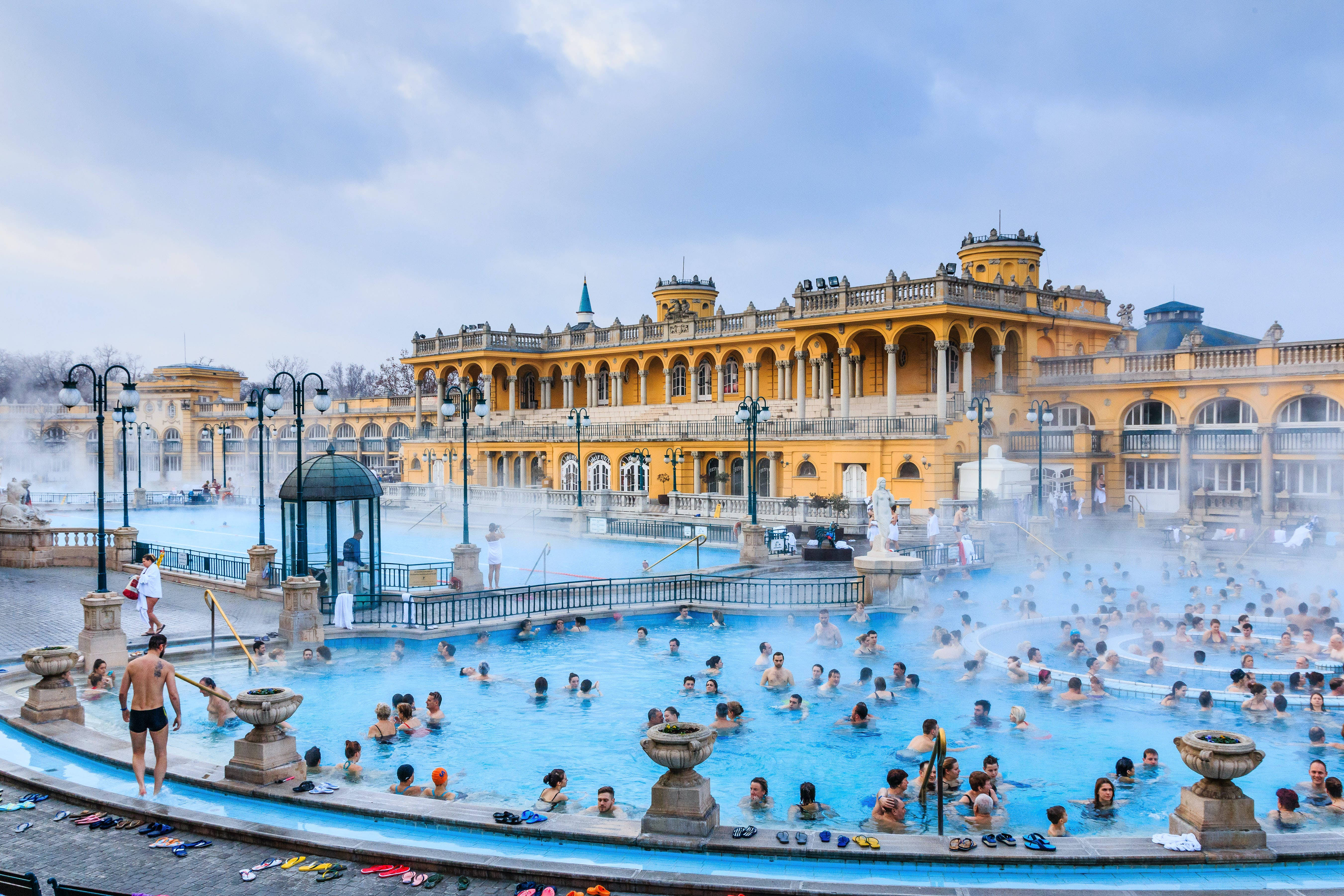 budapest 桌布 壁紙 — 蒸氣從熱鬧的戶外溫泉池中升起，人們在陰天時於一座宏偉的黃色新古典主義建築附近放鬆身心。