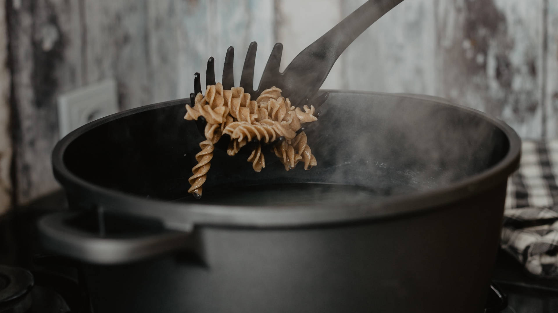 Pasta 桌布 壁紙 — 蒸氣從黑色鍋中升起，叉子舉起煮熟的螺絲粉，背景是質樸風化的牆壁。