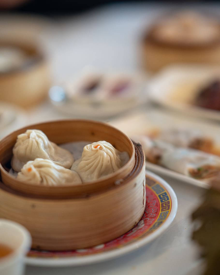 steam 桌布 壁紙 — 蒸氣騰騰的小籠包放在竹蒸籠裡，置於花紋盤上，背景有模糊的點心菜餚。