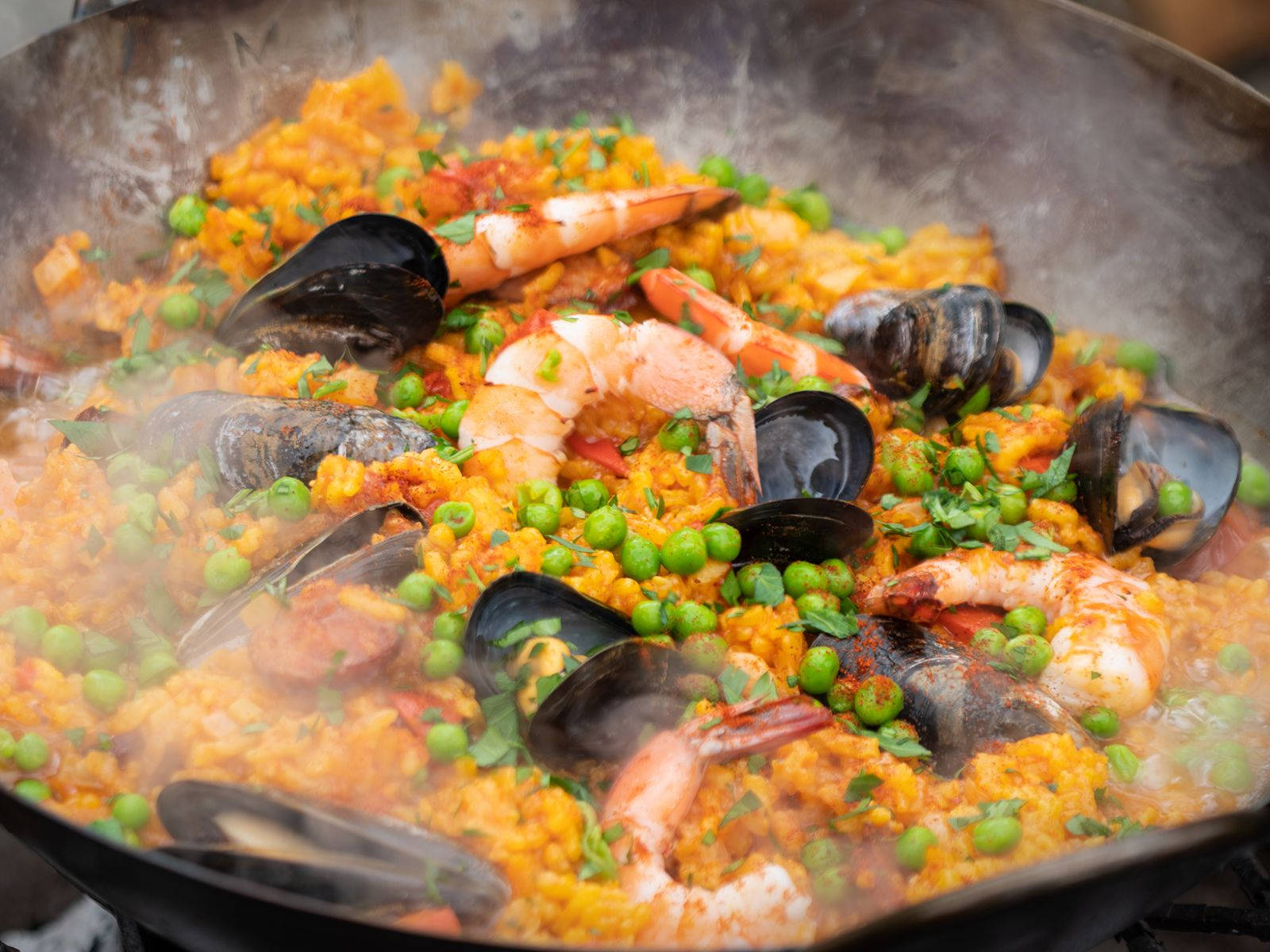 shrimp 桌布 壁紙 — 蒸氣騰騰的海鮮西班牙大鍋飯在黑色平底鍋中滋滋作響，配有蝦、青口、豌豆和香草。