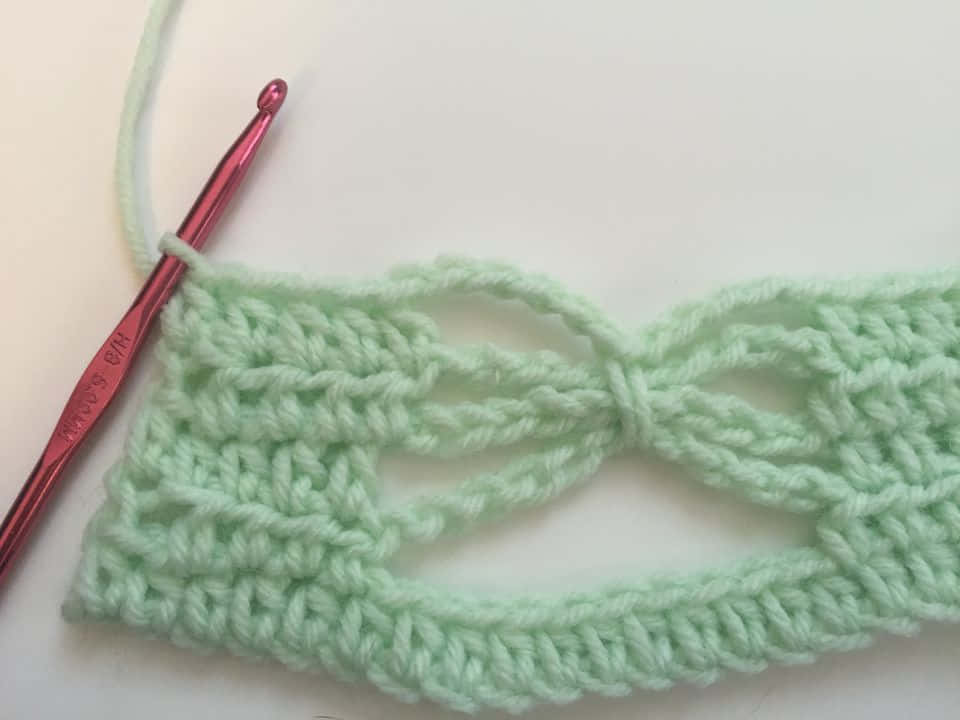 Butterfly Stitch 桌布 壁紙 — 薄荷綠色鉤針編織布料的特寫，帶有蝴蝶結圖案，粉紅色鉤針放在純白色表面上。