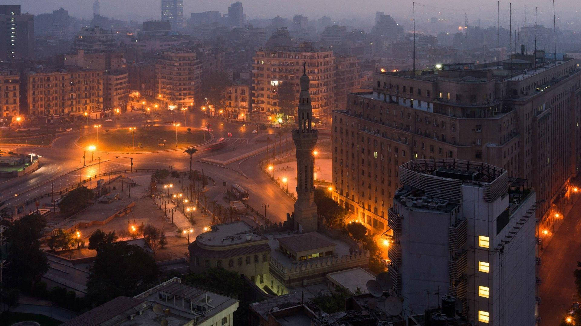 Cairo 桌布 壁紙 — 薄霧籠罩的黃昏城市景觀，街燈照亮了一個圓環和一座高聳的紀念碑，周圍是密集的城市建築。