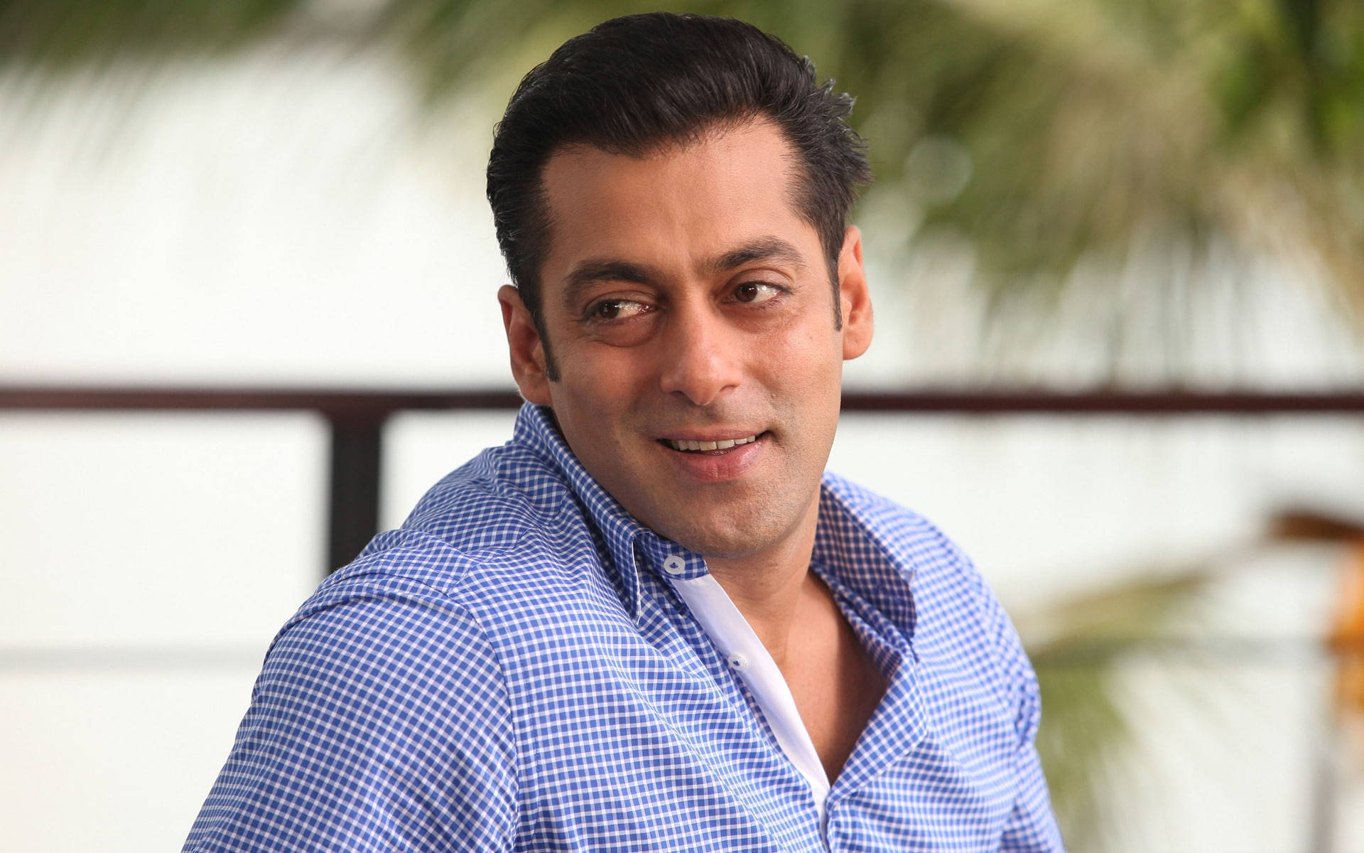 Salman Khan 桌布 壁紙 — 薩爾曼·汗穿著藍色格紋襯衫，在模糊的熱帶背景下溫暖地微笑。