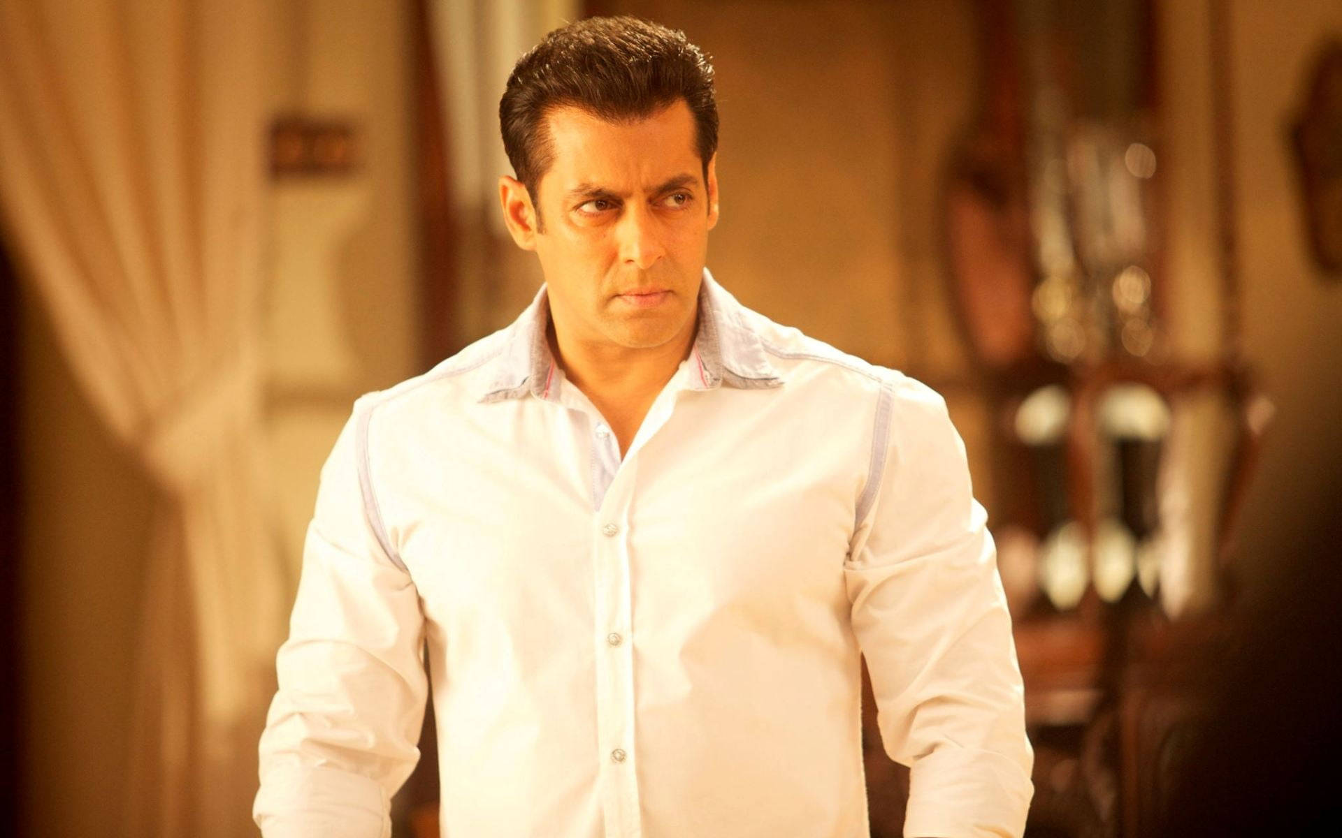 Salman Khan 桌布 壁紙 — 薩爾曼·汗站在一個光線溫暖的房間裡，穿著一件挺括的白襯衫，表情嚴肅。