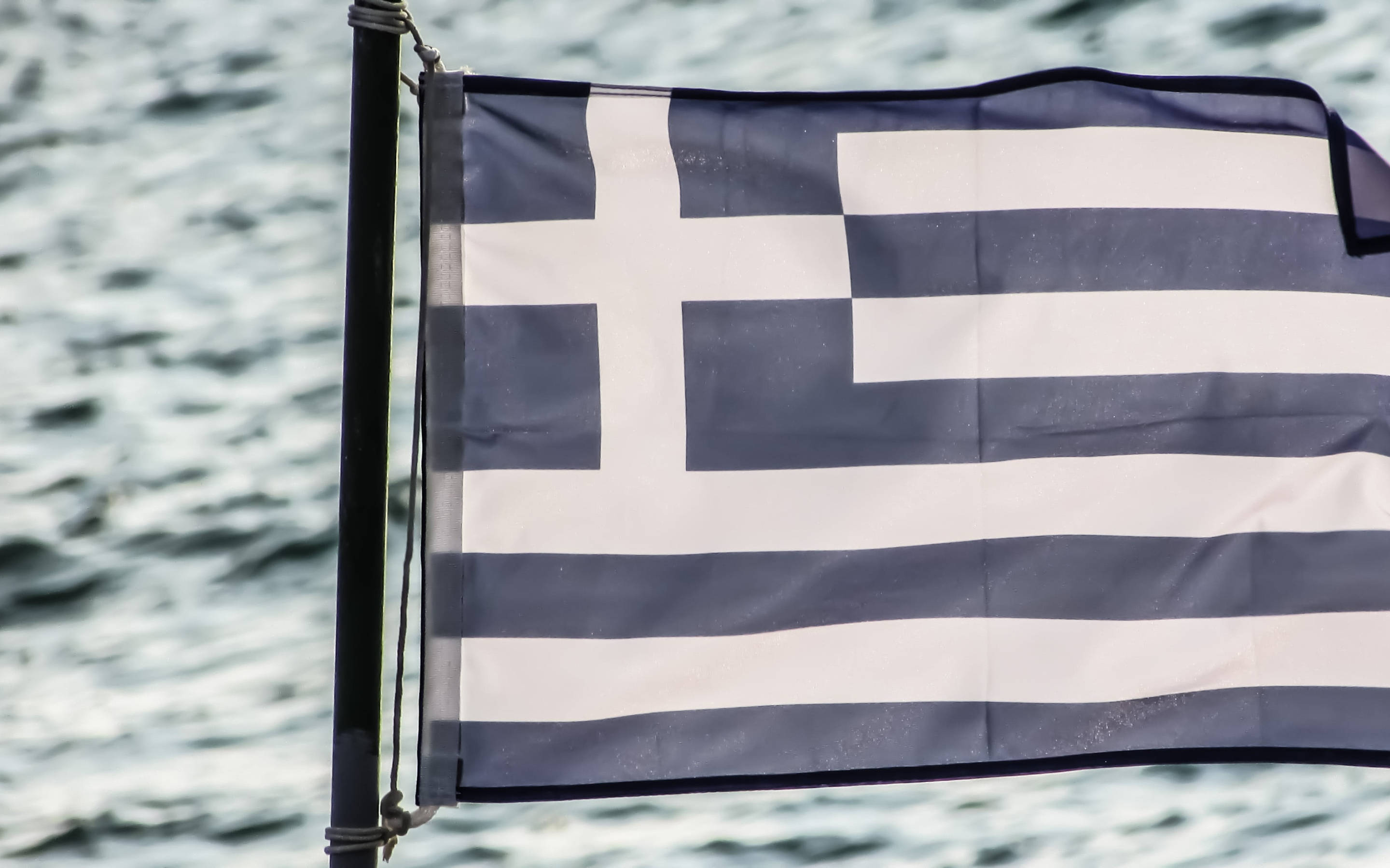 greek 桌布 壁紙 — 藍白條紋和十字架的希臘國旗在桿子上飄揚，背景是波光粼粼的海水。