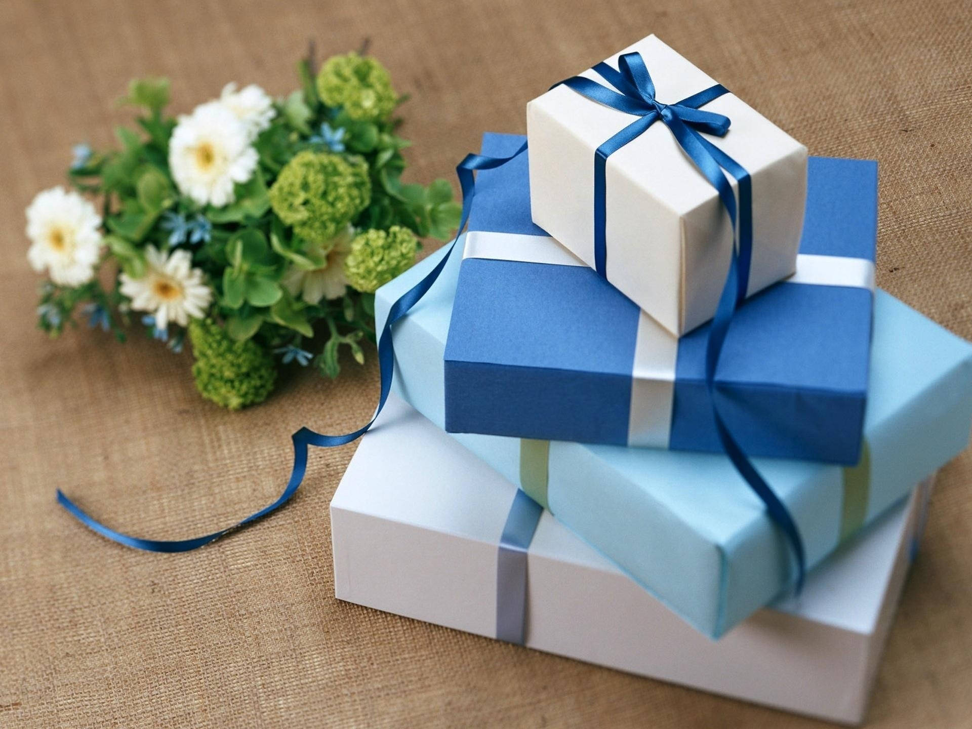 Gift 桌布 壁紙 — 藍白相間的禮品盒堆疊在一起，用絲帶繫著，旁邊放著一束白色鮮花，放在粗麻布上。