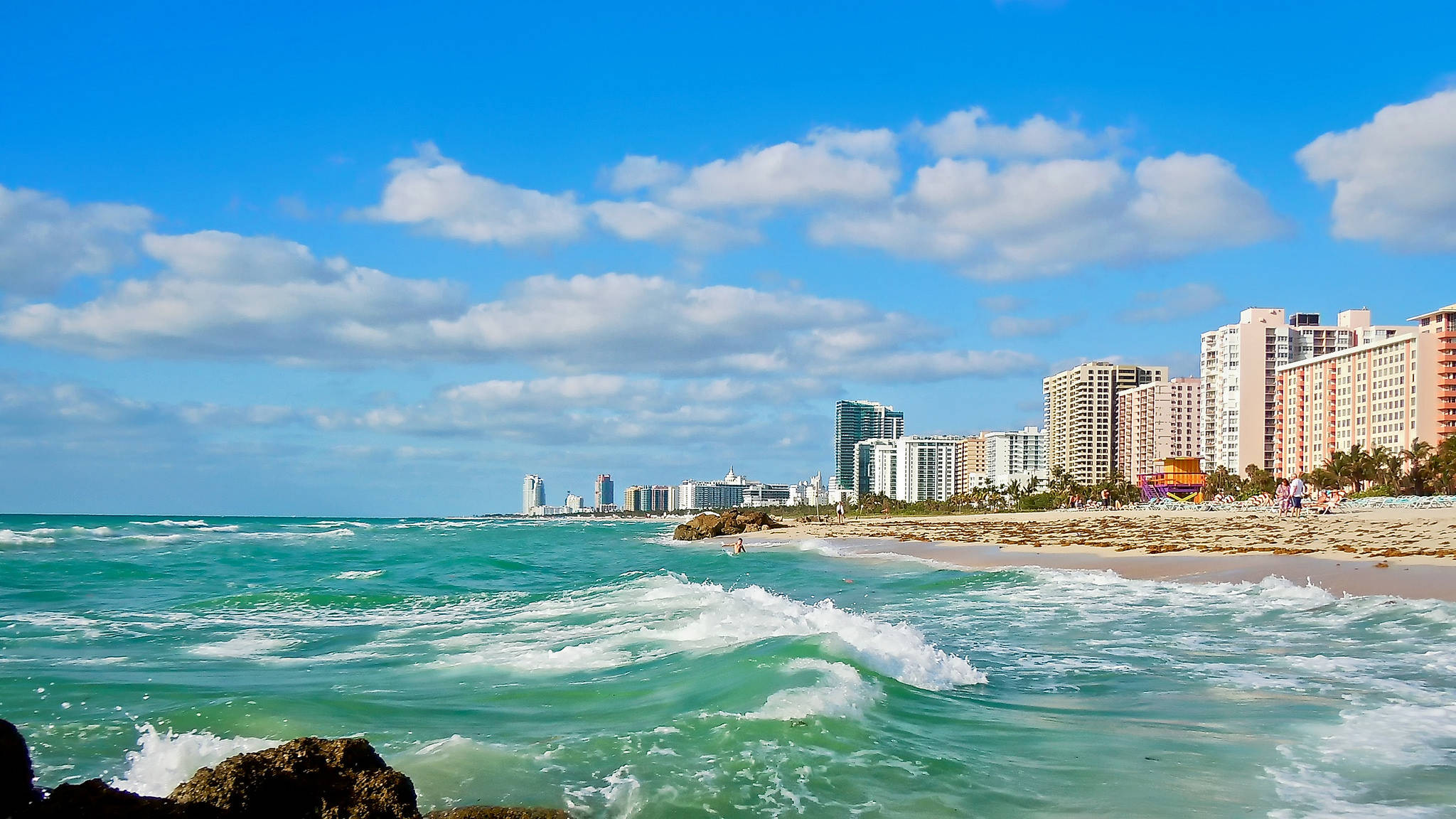 Miami 桌布 壁紙 — 藍綠色海浪拍打著沙灘，沙灘兩旁高聳的酒店林立，天空湛藍，白雲朵朵。