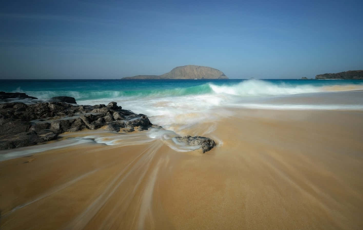 Beautiful Beach 桌布 壁紙 — 藍綠色海浪拍打金色沙灘，旁邊有深色岩石，遠處島嶼在清澈藍天下。