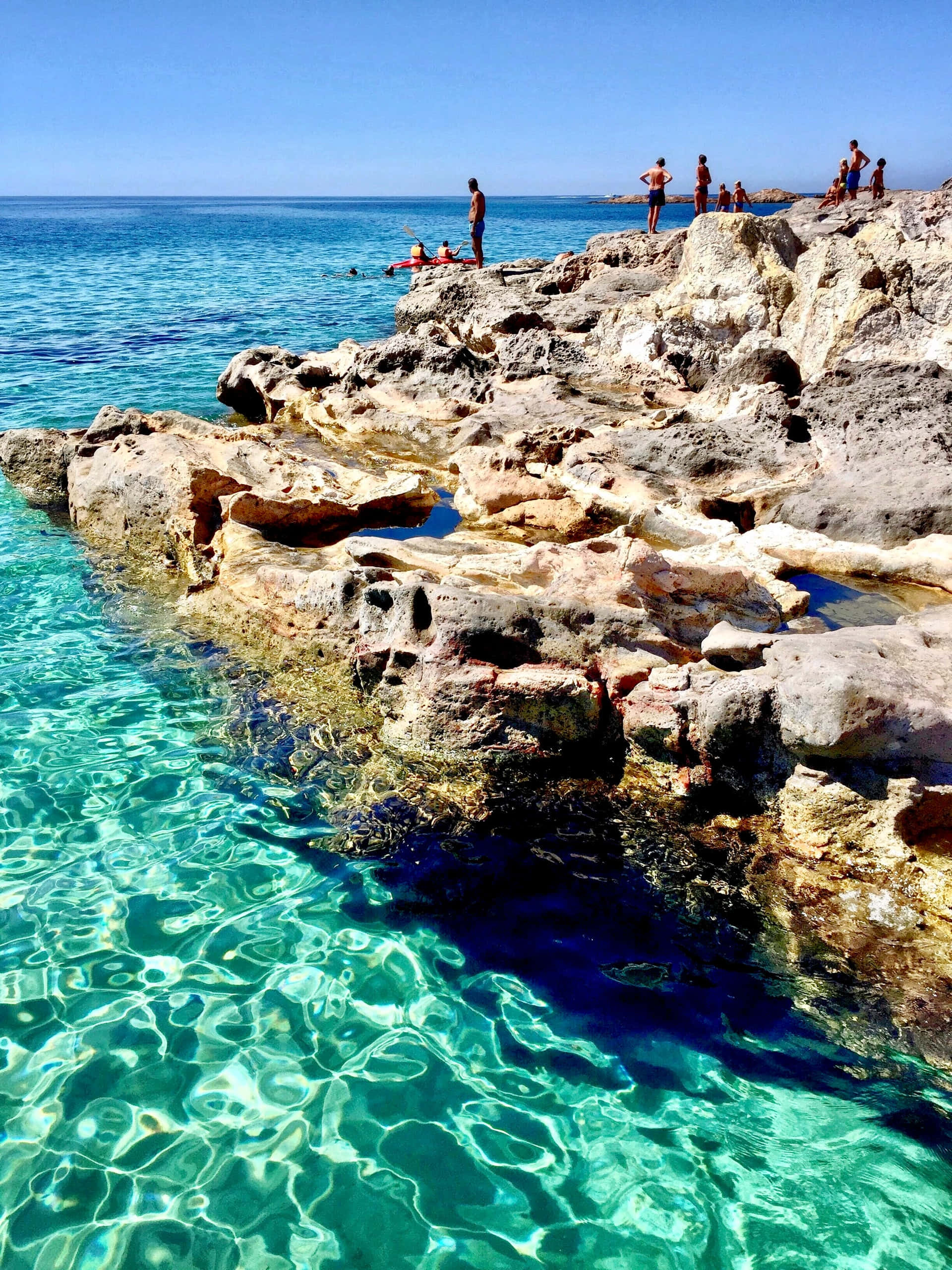 Italian Beach 桌布 壁紙 — 藍綠色的海水在陽光照耀的岩石懸崖周圍泛起漣漪，人們在晴朗的藍天下放鬆和游泳。