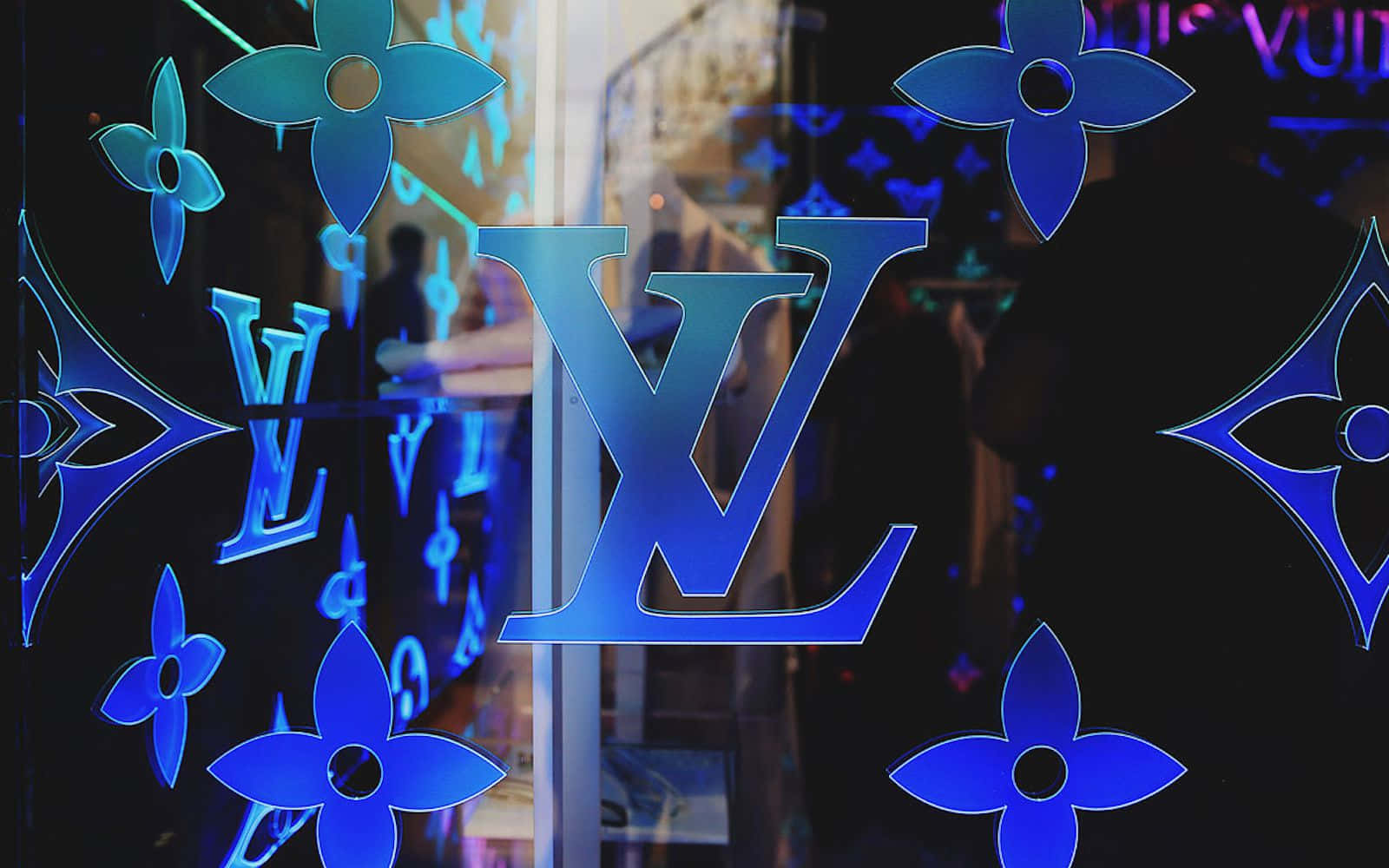 Louis Vuitton Pattern 桌布 壁紙 — 藍色 Louis Vuitton 字母組合圖案在玻璃店面上發光，帶有模糊的室內倒影。