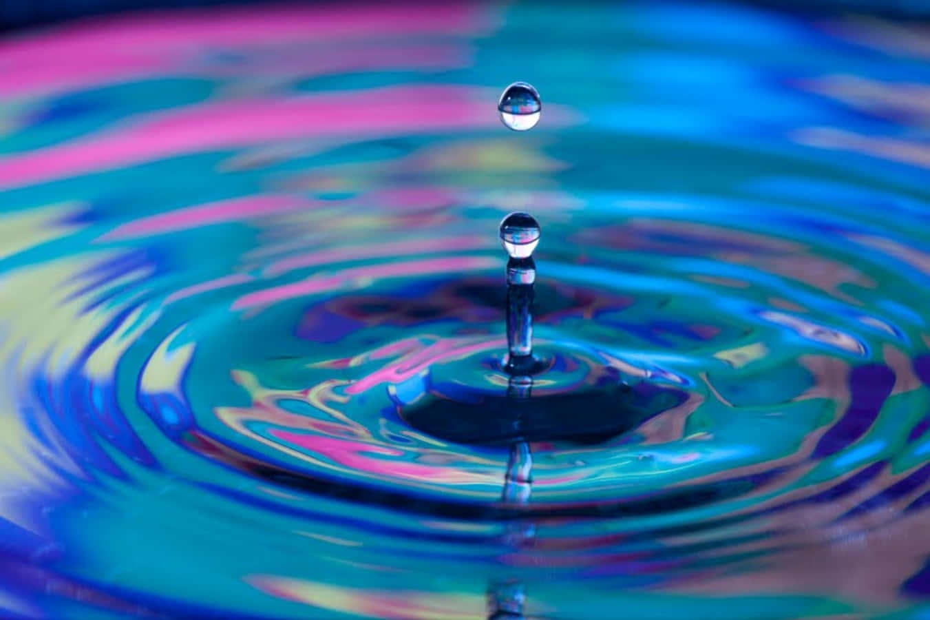 Water Drop 桌布 壁紙 — 藍色、粉紅色和黃色的鮮豔水波紋從懸浮的水滴向外擴散，在凝固的瞬間捕捉光線。