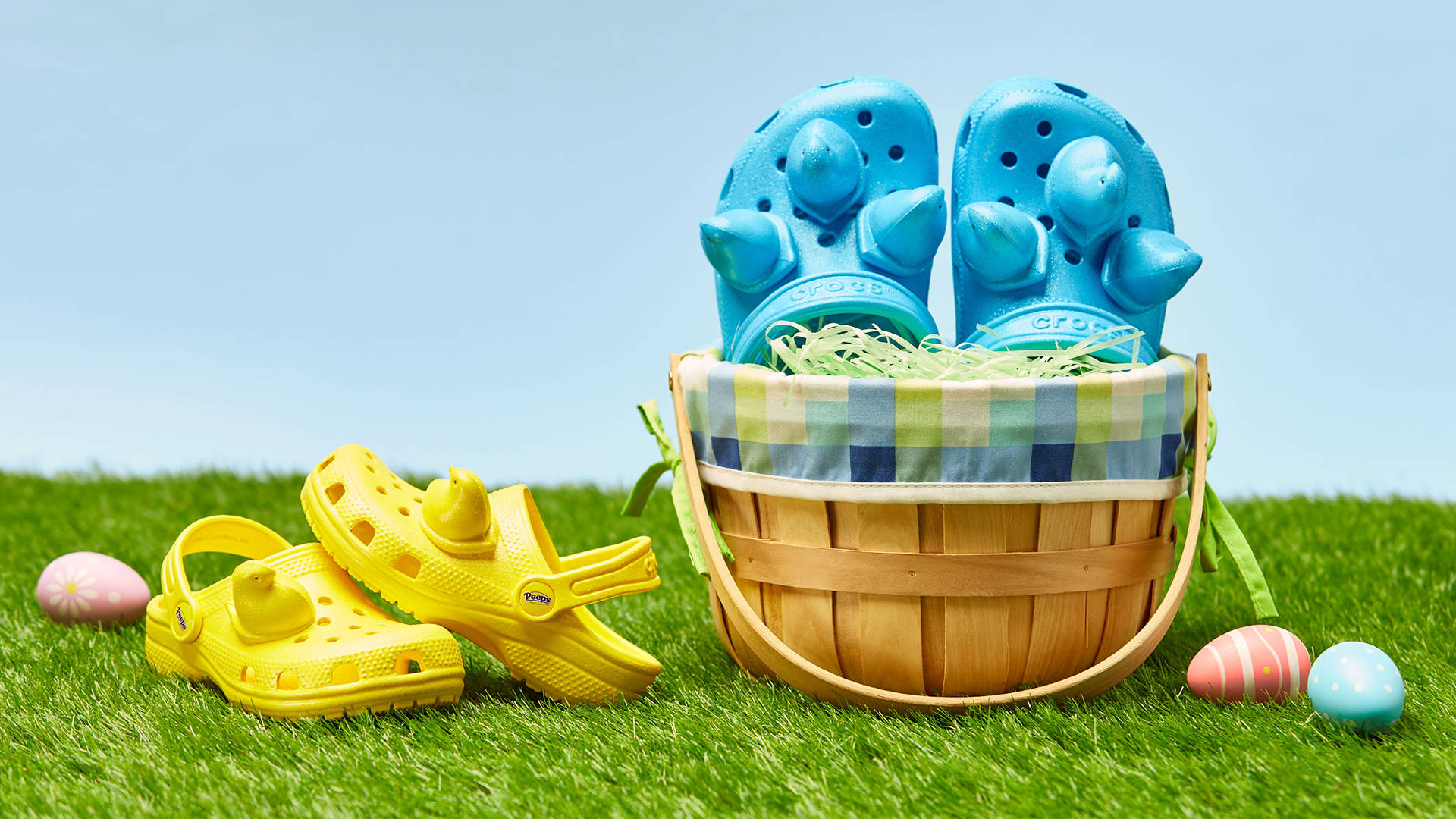 Crocs 桌布 壁紙 — 藍色和黃色的Crocs涼鞋放在一個編織籃子裡，裡面有復活節彩蛋，放在充滿活力的綠色草地上，天空晴朗。