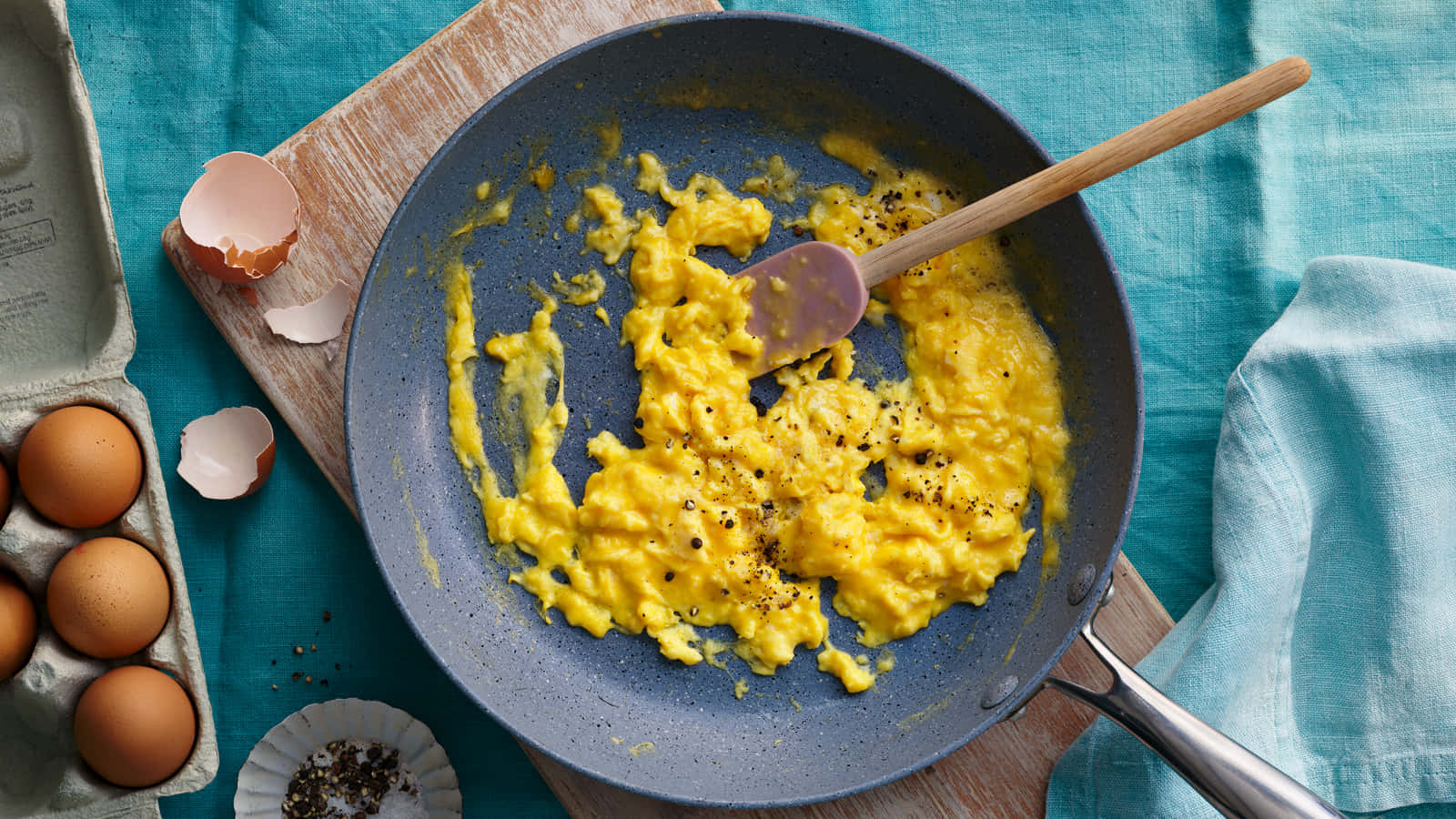 Scrambled Egg 桌布 壁紙 — 藍色斑點平底鍋中的金黃色炒蛋配黑胡椒，放在木板上，旁邊有雞蛋盒和碎蛋殼。