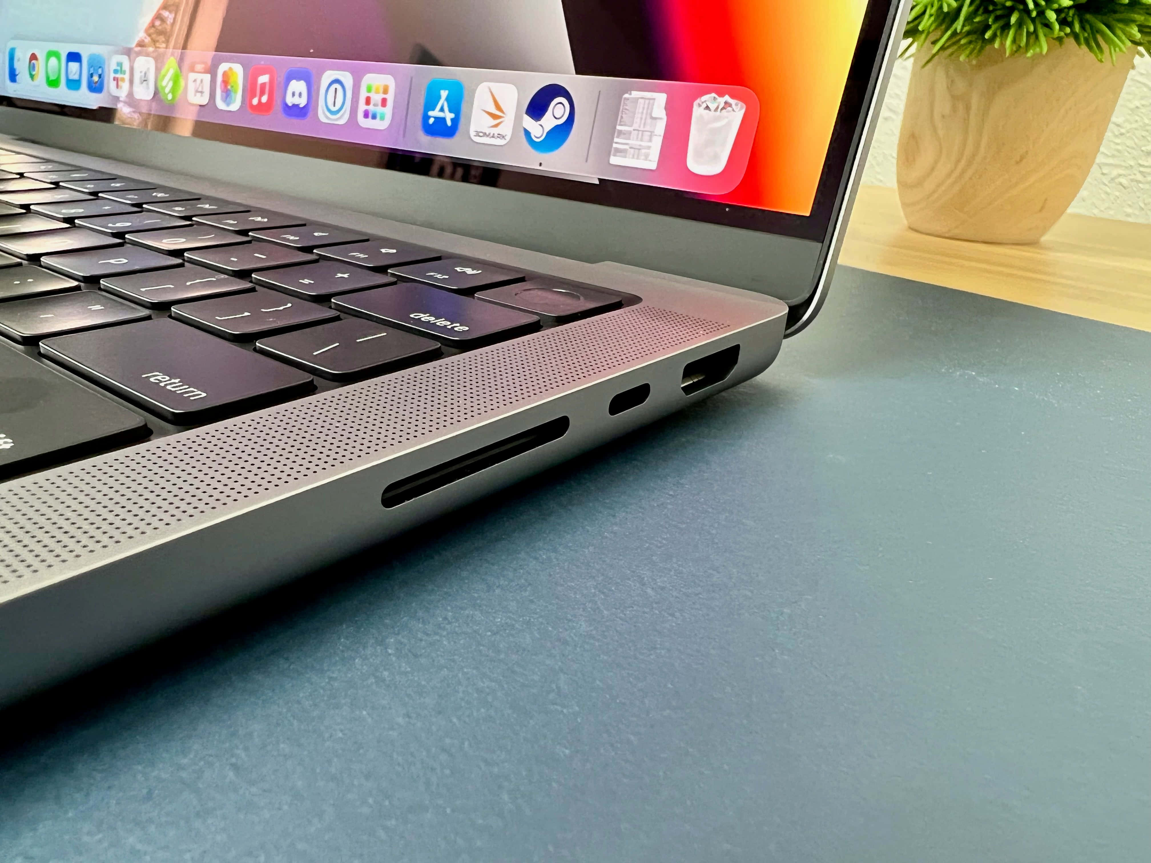 MacBook 動態圖片 桌布 壁紙 — 藍色書桌上時尚銀色筆記型電腦的特寫，附近有盆栽植物，螢幕上顯示色彩繽紛的應用程式圖示。