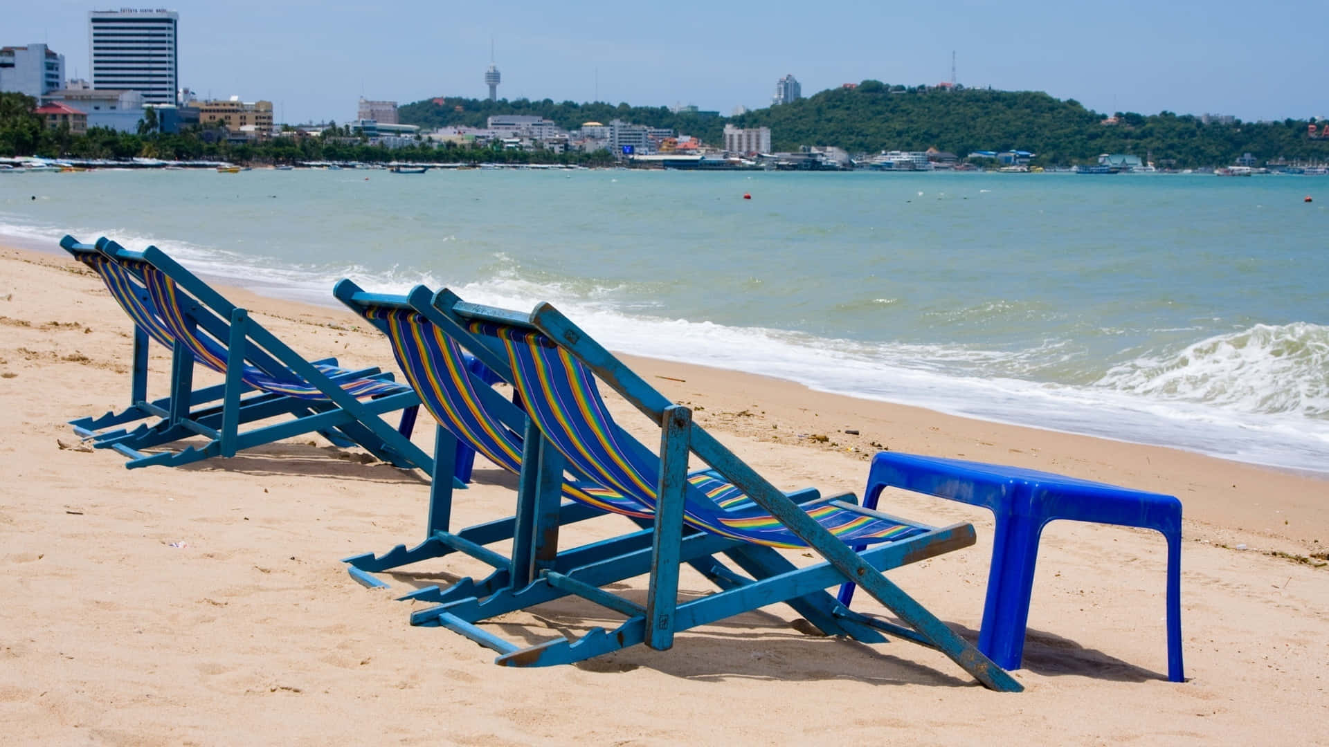 Beach Sunbed 桌布 壁紙 — 藍色條紋沙灘椅空置在沙灘上，面向碧綠海浪與遠處城市景觀，天空晴朗。