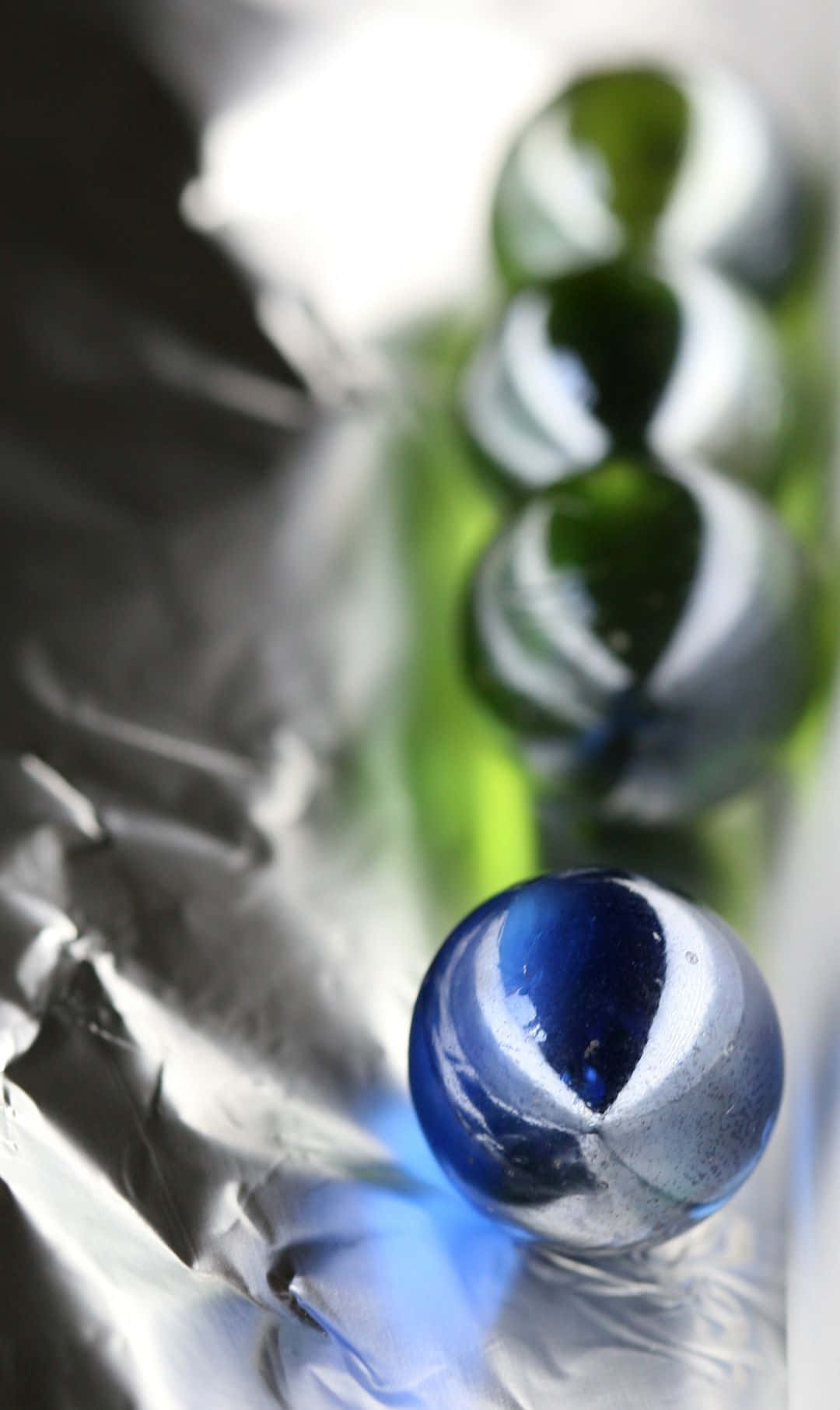1080x1812 桌布 壁紙 — 藍色玻璃彈珠的特寫，在皺褶的銀色錫箔紙上反射光線，背景有綠色玻璃球模糊。