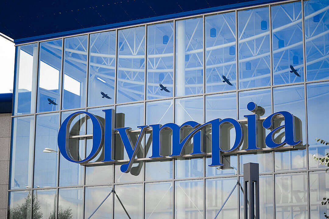 標誌 桌布 壁紙 — 藍色的'Olympia'標誌在玻璃建築物外牆上閃耀，反射著天空和飛翔的鳥兒。