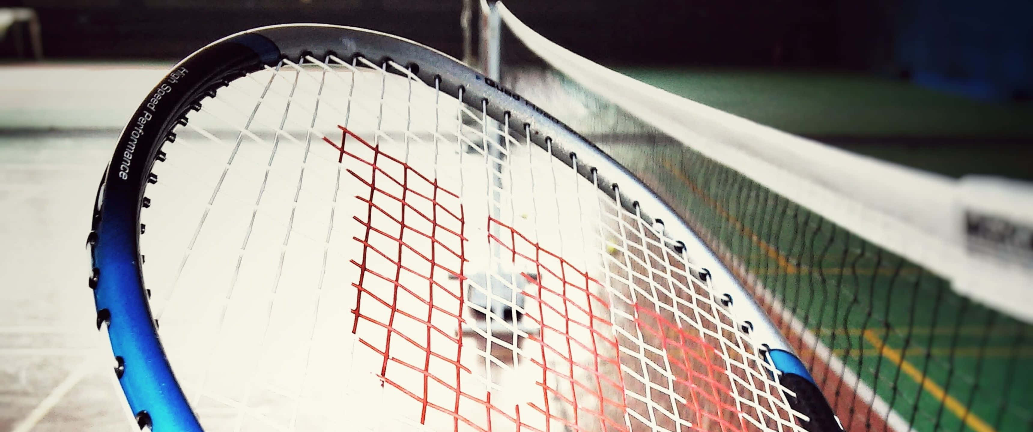 3440x1440p Badminton 桌布 壁紙 — 藍色網球拍頭的近距離特寫，帶有紅色和白色線，背景是模糊的網球場網和綠色表面。
