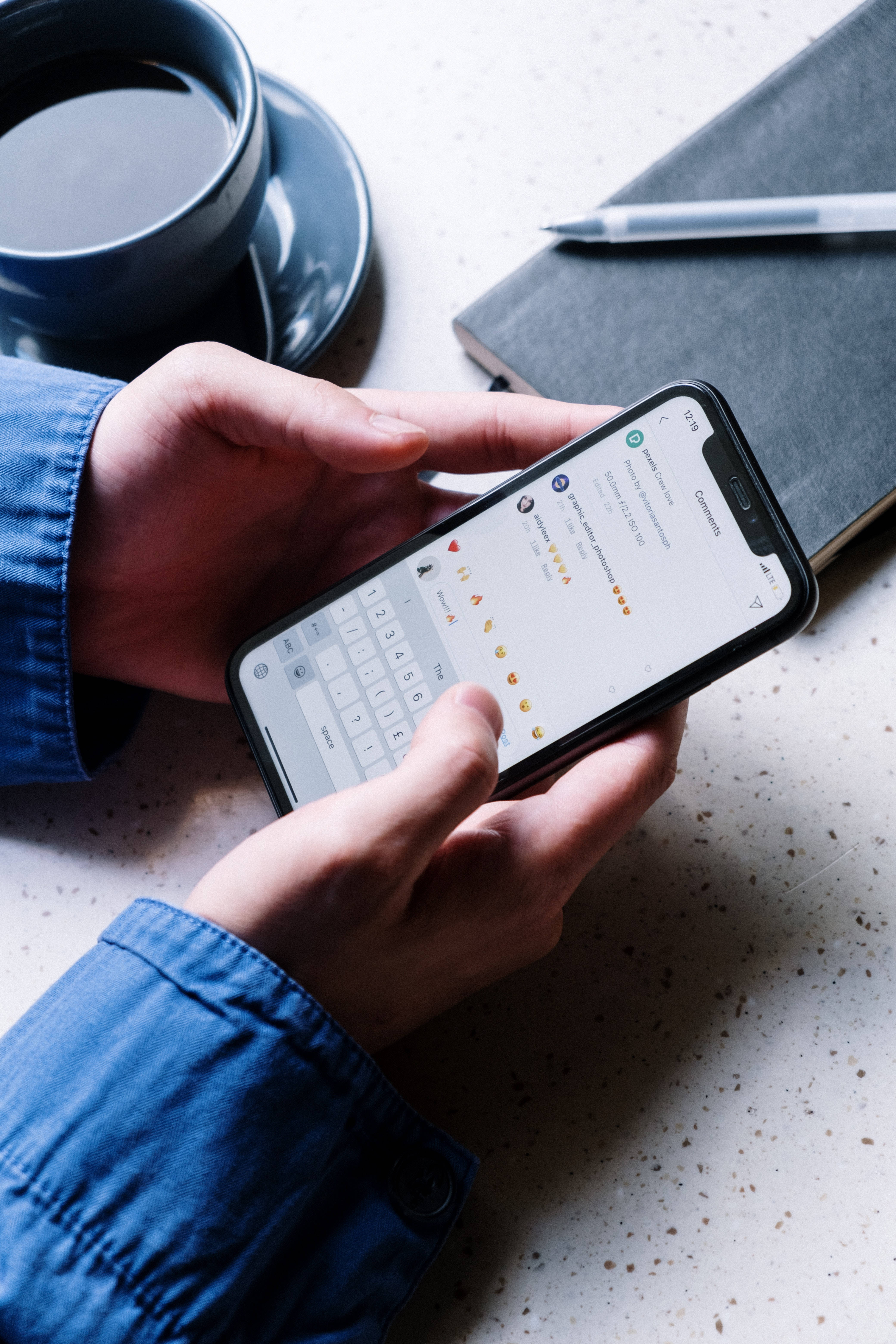 酷炫iPhone 桌布 壁紙 — 藍色袖子的手在斑點檯面上，於咖啡杯和筆記本旁邊的智慧型手機上打字訊息。
