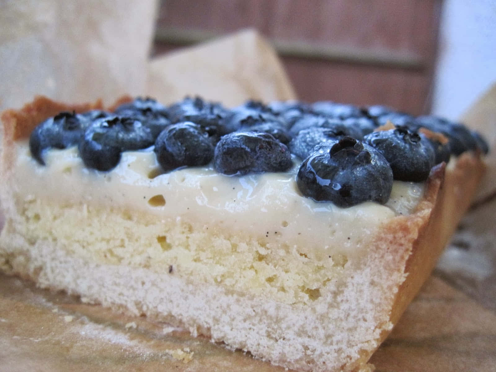 Blueberries Tart 桌布 壁紙 — 藍莓塔一片的特寫，有奶油餡料和金黃色塔皮，放在烘焙紙上。