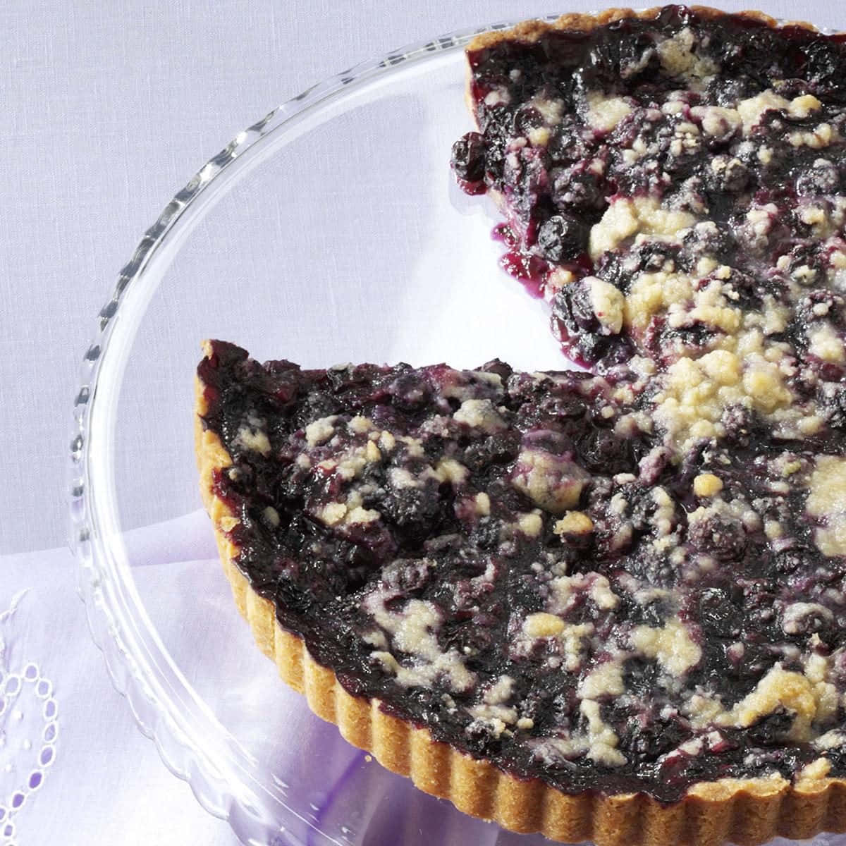 Blueberries Tart 桌布 壁紙 — 藍莓金寶塔近距離特寫，塔邊有凹槽紋路，放在玻璃盤上，襯以淺紫色布料。