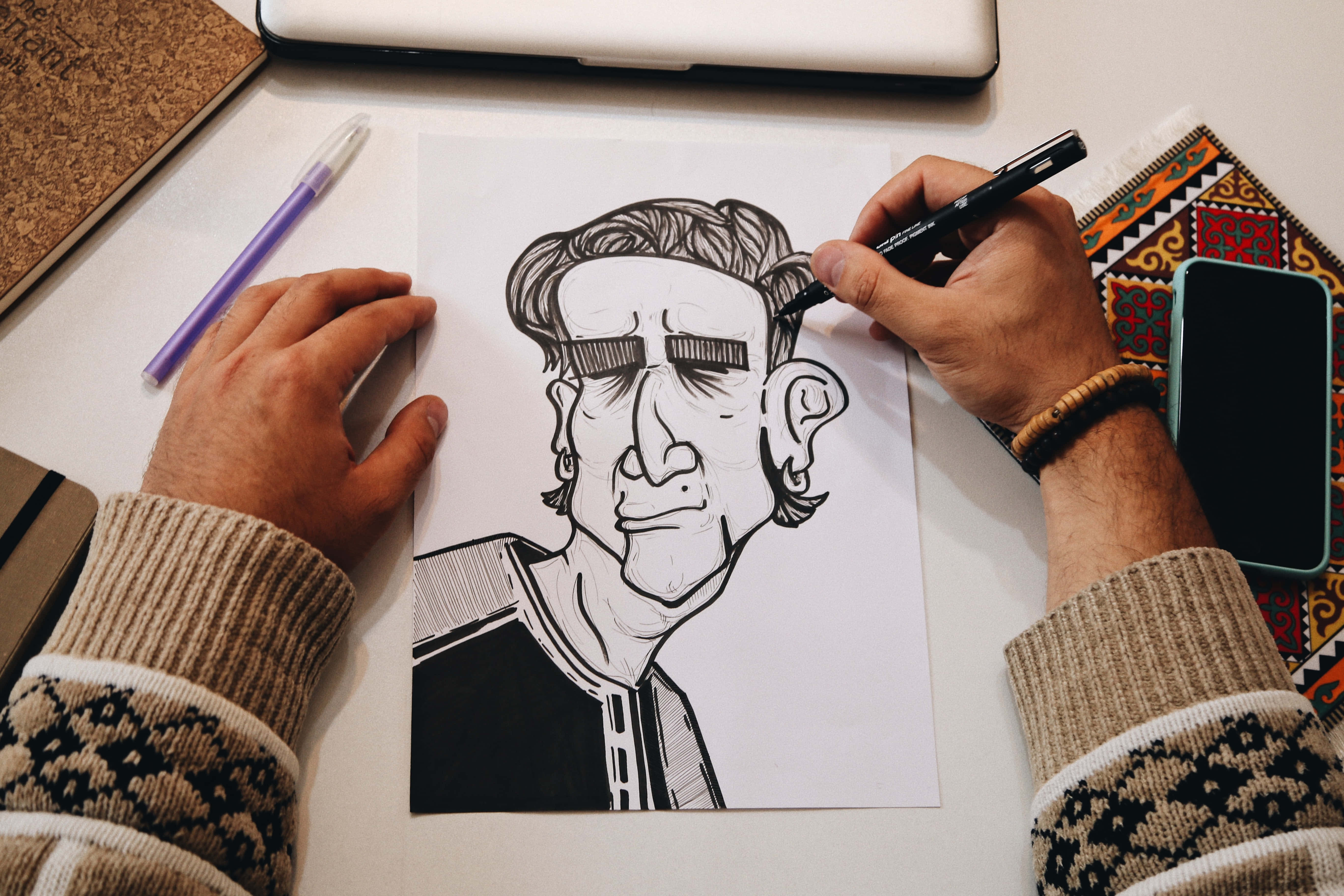 Drawing 桌布 壁紙 — 藝術家的手在白色桌子上，被藝術用品包圍，用馬克筆繪製詳細的黑白漫畫肖像。