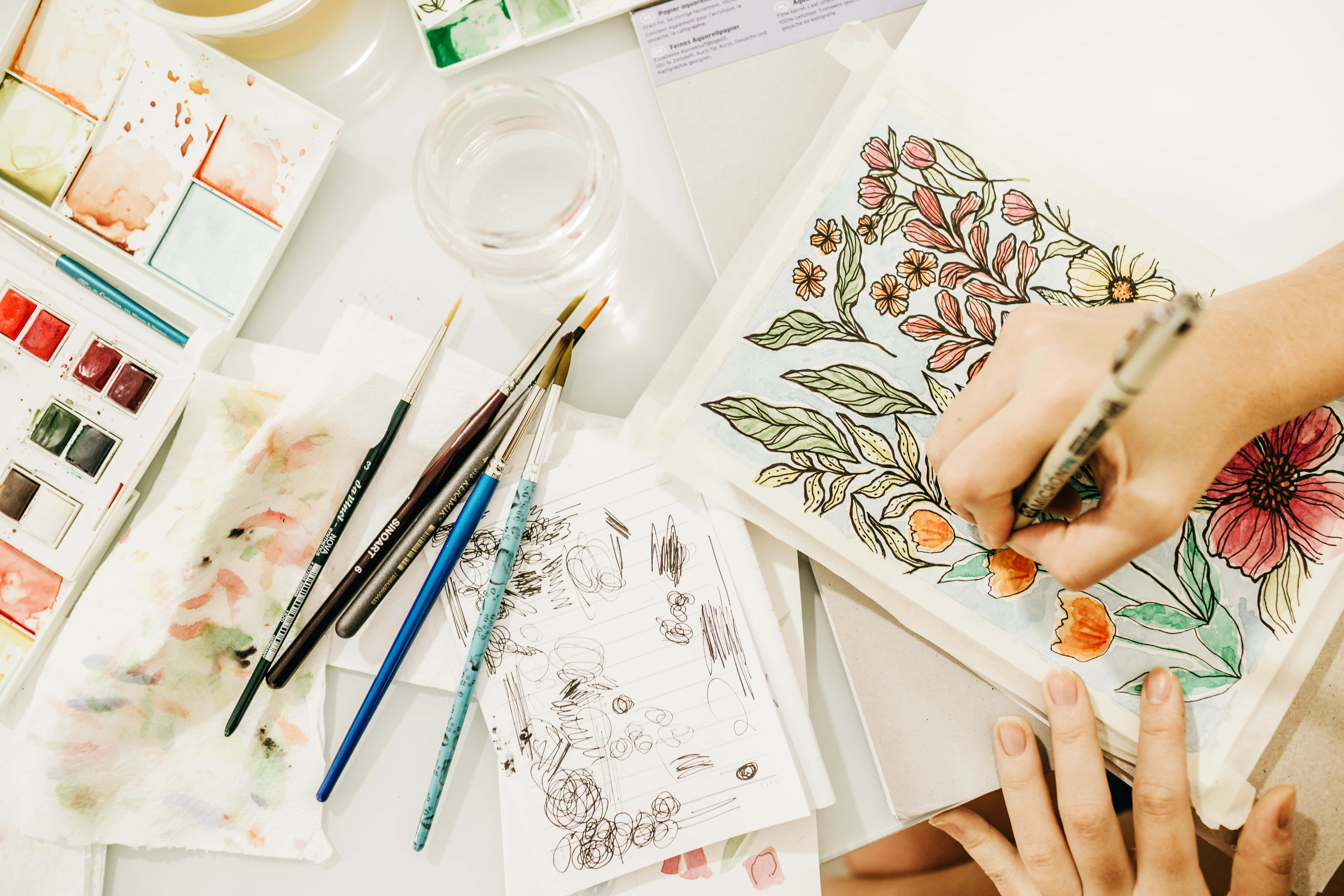 Drawing 桌布 壁紙 — 藝術家的手用畫筆為花卉插圖上色，周圍是水彩顏料和畫筆，放在白色桌子上。