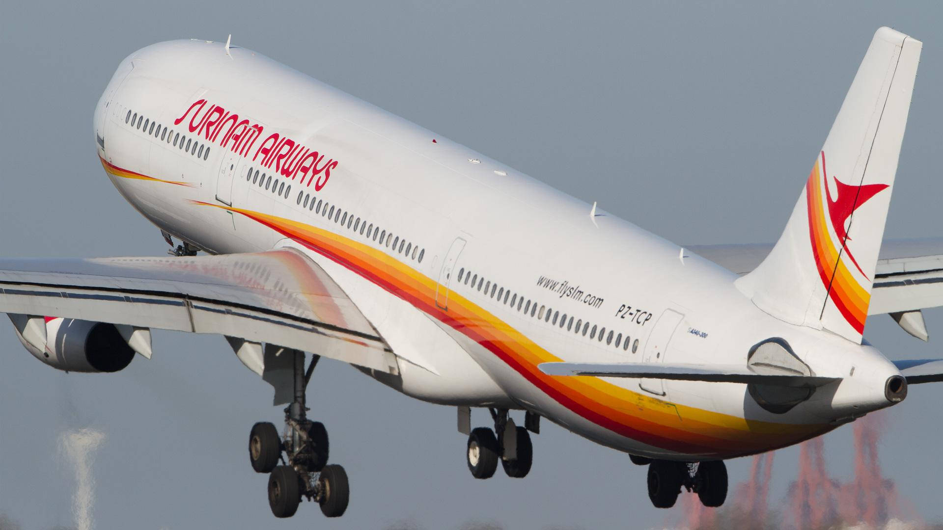 Suriname 桌布 壁紙 — 蘇利南航空一架紅橙黃色塗裝的空中巴士A340在起飛時急轉，背景是朦朧的天空。