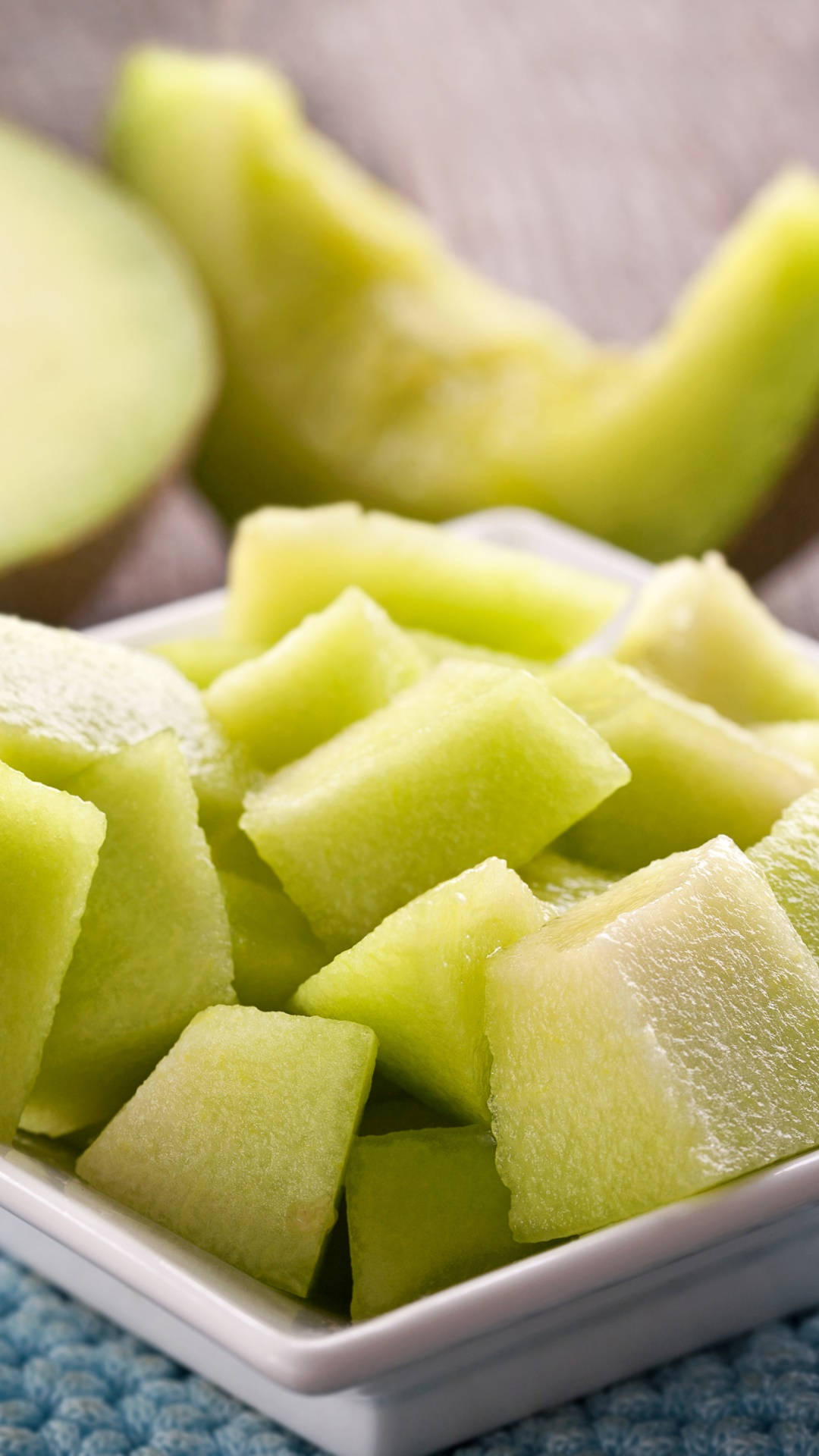 Fruits 桌布 壁紙 — 蜜瓜丁在白色碗中閃閃發光，置於藍色紋理布上，背景有半個蜜瓜模糊可見。