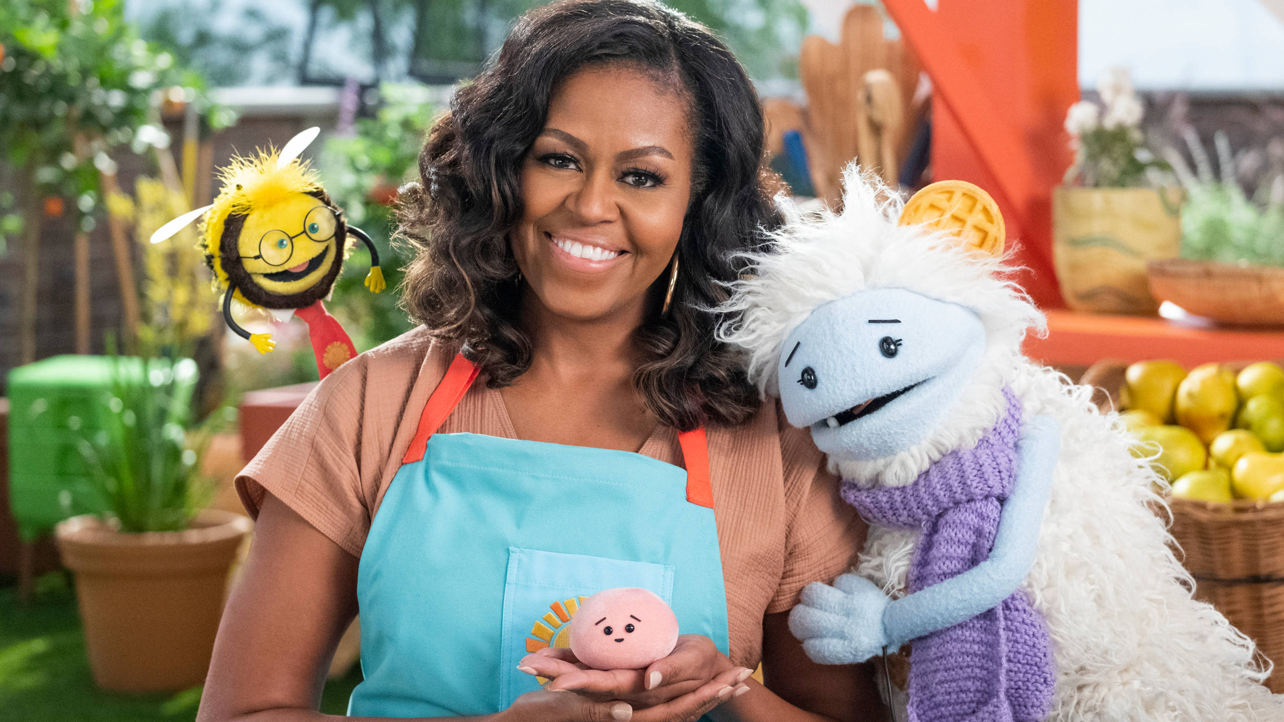 Michelle 桌布 壁紙 — 蜜雪兒·歐巴馬在充滿活力的花園中溫暖地微笑，手持一個粉紅色木偶，旁邊有一隻蜜蜂和一個毛茸茸的藍色木偶。