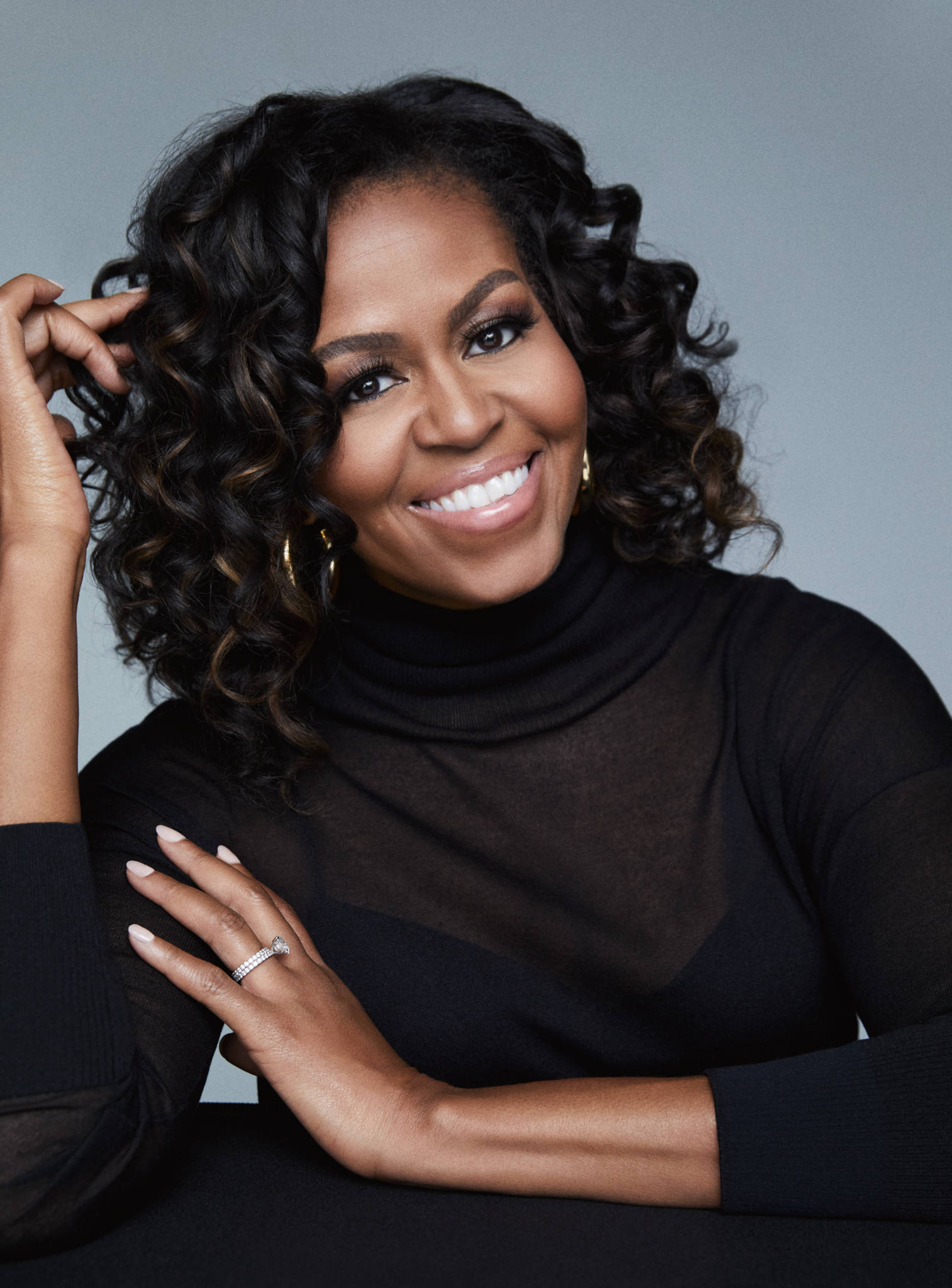 Michelle 桌布 壁紙 — 蜜雪兒·歐巴馬在柔和的灰色攝影棚背景下，雙手靠近捲髮，溫暖地微笑。