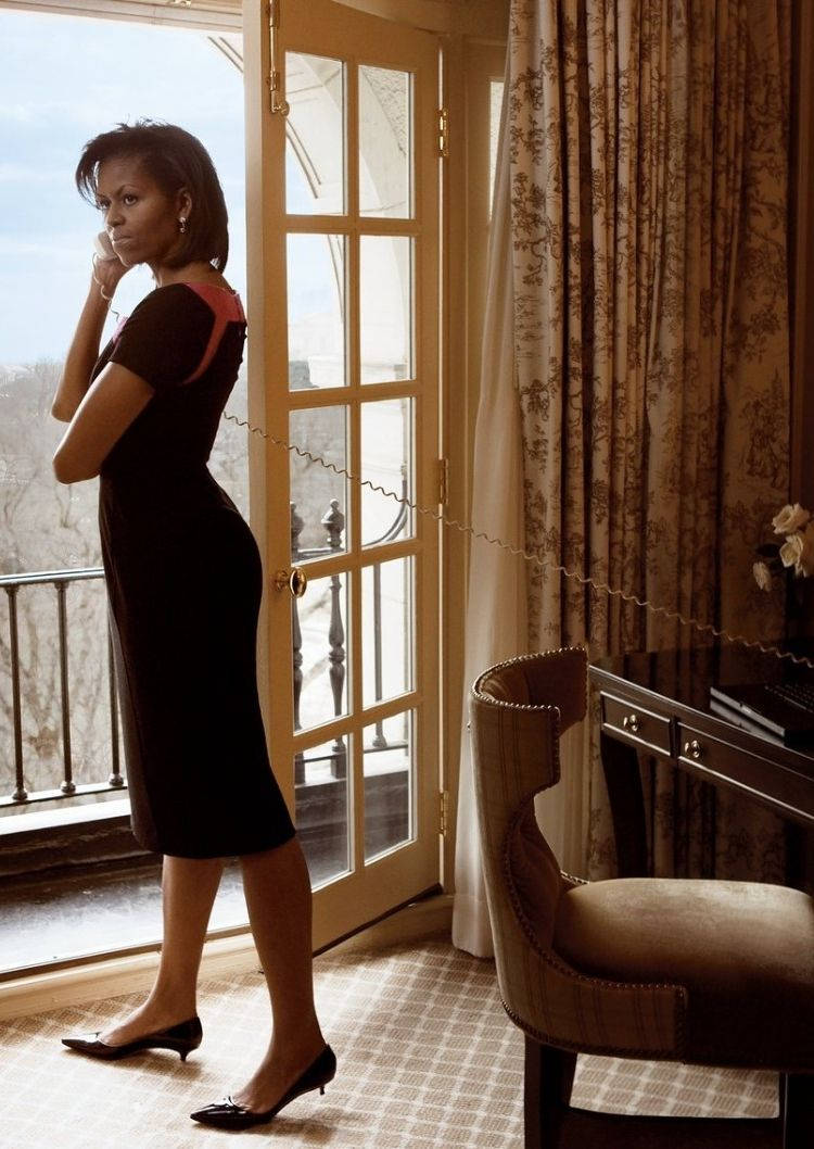 Michelle 桌布 壁紙 — 蜜雪兒·歐巴馬站在陽光充足的房間裡的玻璃門旁，身穿黑色連衣裙和高跟鞋，若有所思地凝視著外面。