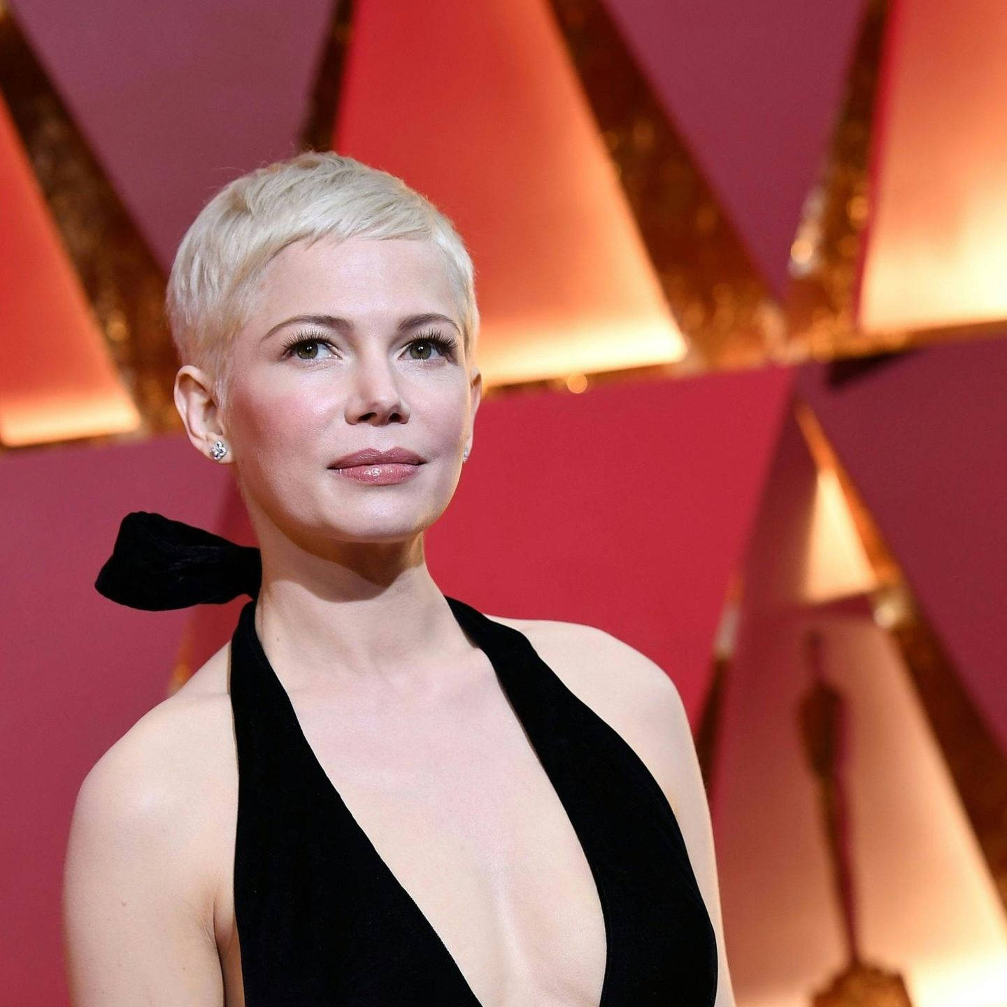 oscars 桌布 壁紙 — 蜜雪兒威廉絲身穿黑色天鵝絨長裙，在發光的幾何紅金背景前優雅擺姿勢。