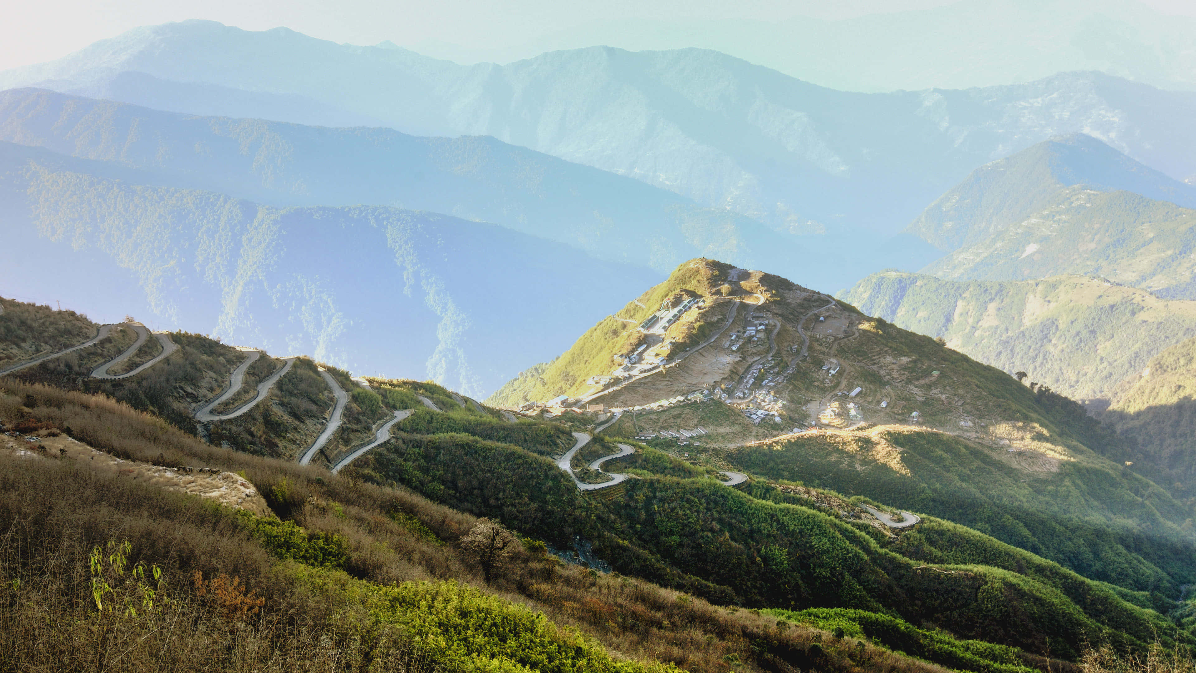 風景 桌布 壁紙 — 蜿蜒的山路在陽光照耀的綠色山丘間蜿蜒，層層疊疊的藍色山峰逐漸消失在朦朧的遠方。
