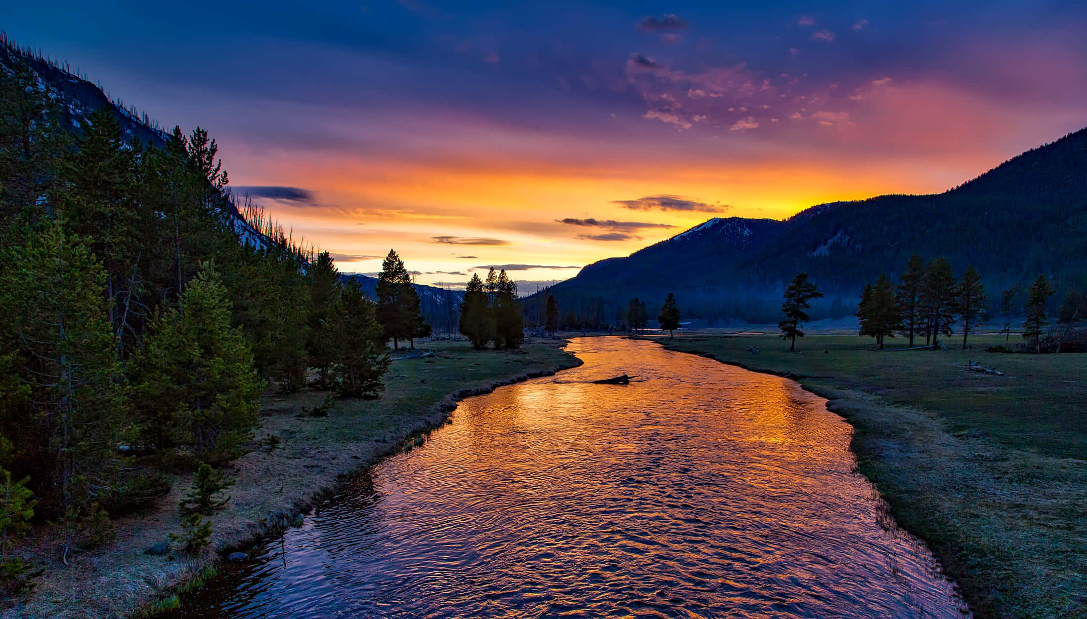 Yellowstone 桌布 壁紙 — 蜿蜒的河流映照出火熱的橙色日落色調，與深紫色的山脈和常綠樹木形成對比。