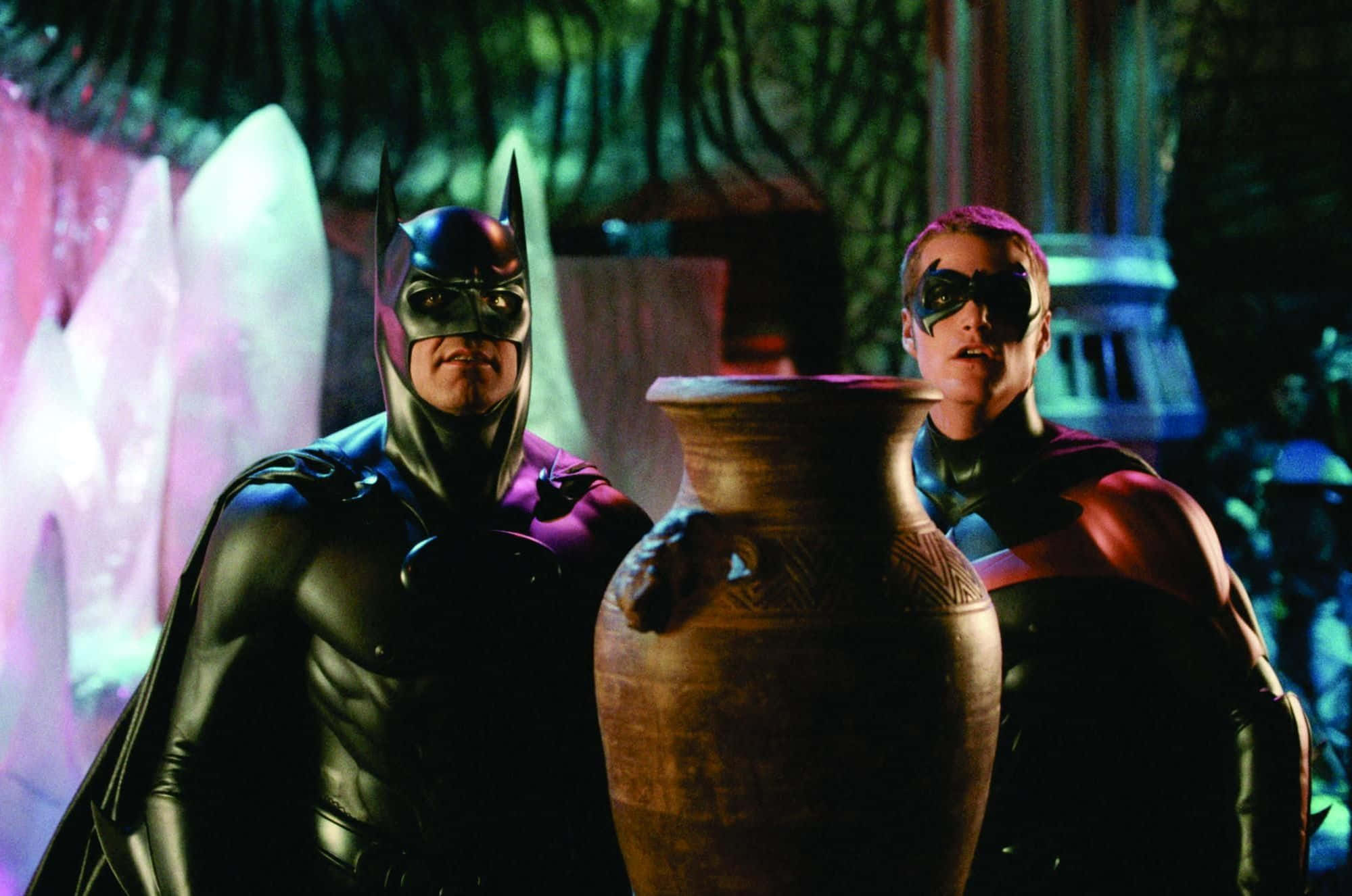 Batman And Robin 桌布 壁紙 — 蝙蝠俠和羅賓站在一個燈光昏暗、神秘的洞穴中，手持一個大型古甕，背景中閃爍著粉紅色的水晶。