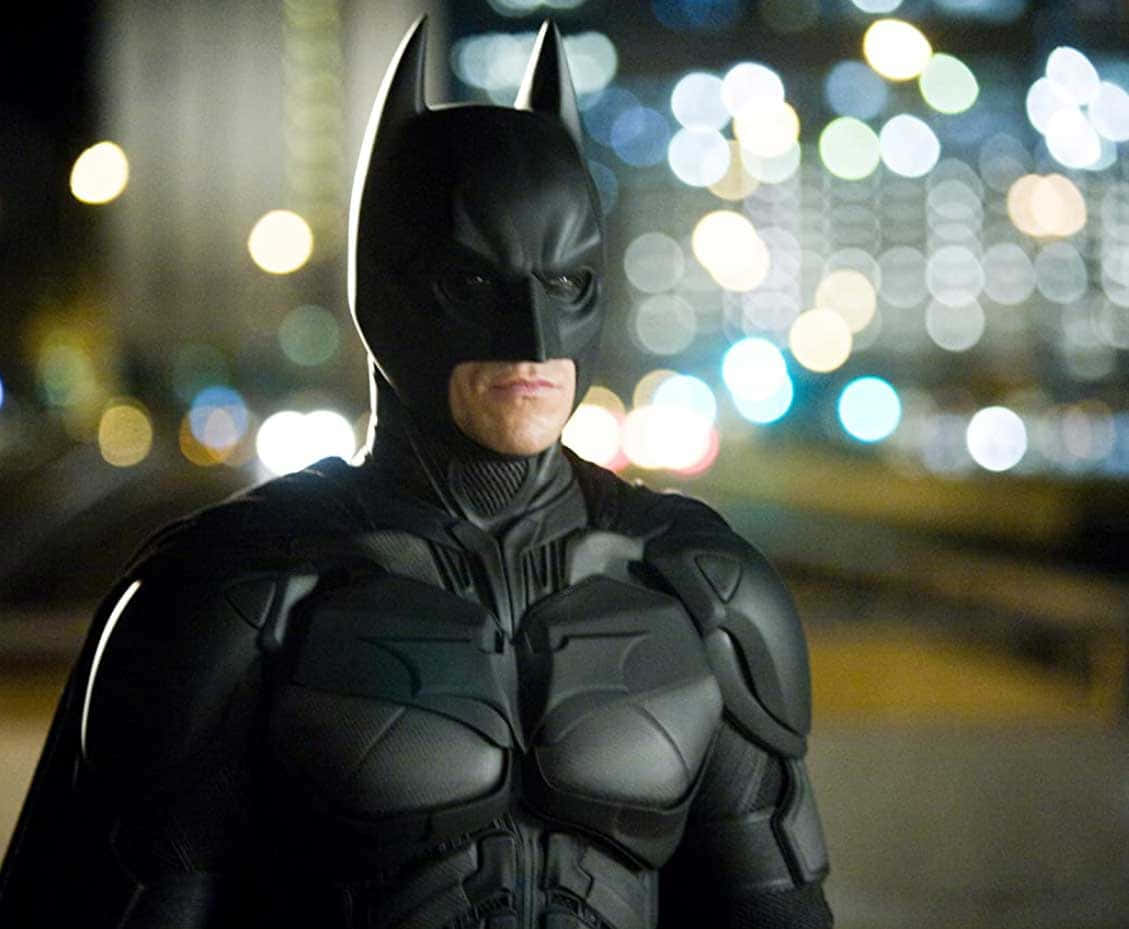 Bat-suit 桌布 壁紙 — 蝙蝠俠在夜晚的黑暗城市景觀中站立，身後有發光的散景燈光。
