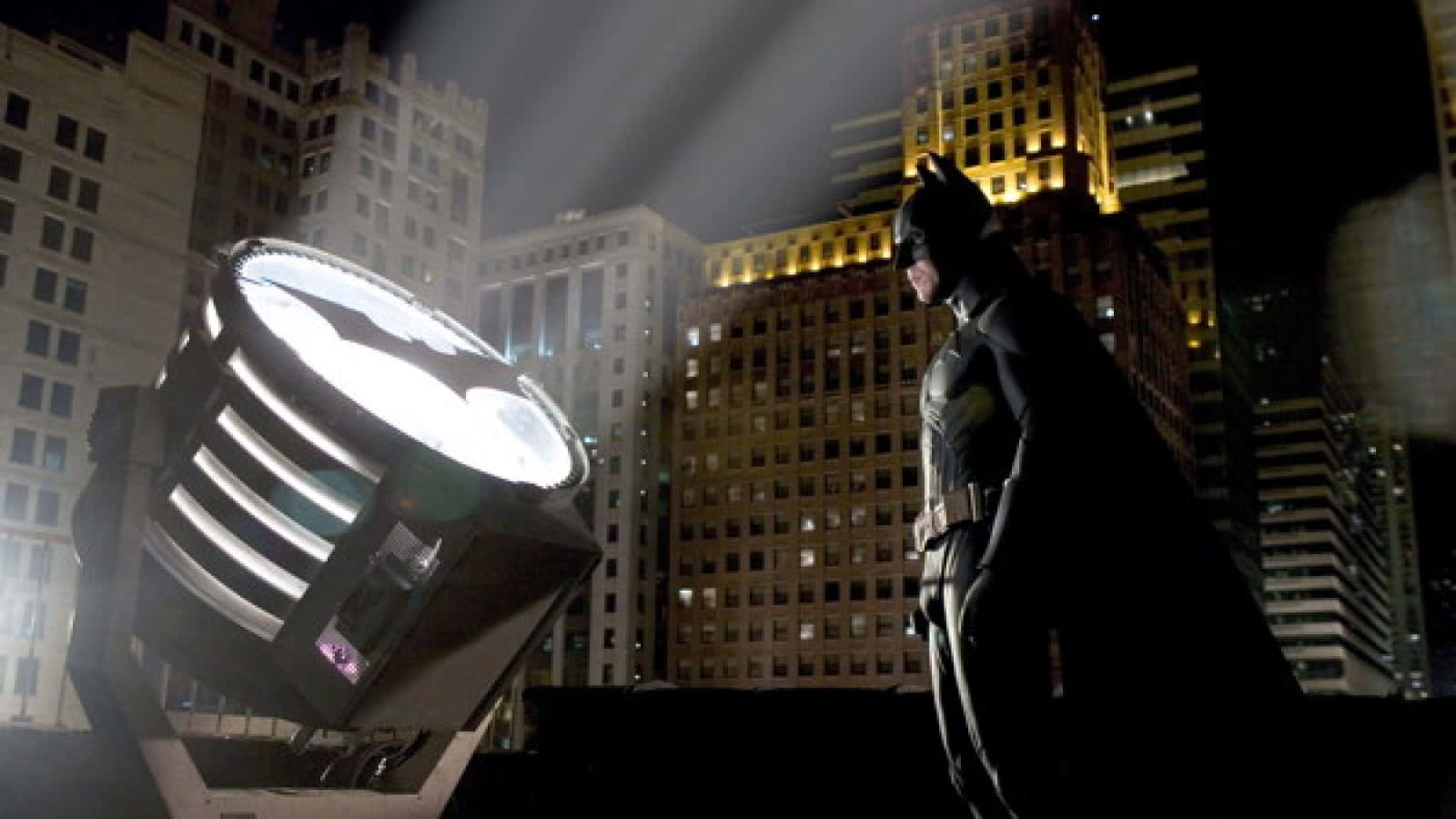 Batman Begins 桌布 壁紙 — 蝙蝠俠在夜間城市天際線上投射的發光蝙蝠信號的襯托下，以剪影姿態站立。