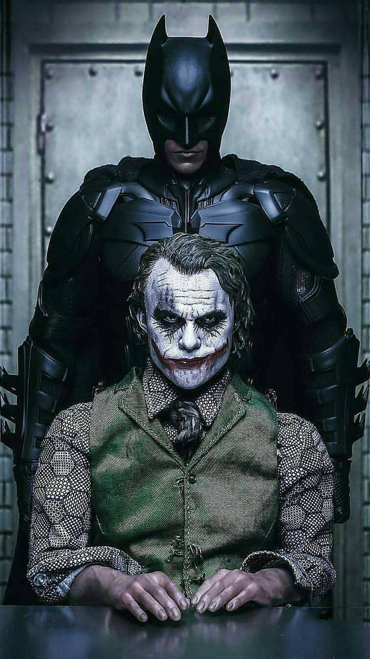 The Batman iPhone 桌布 壁紙 — 蝙蝠俠在昏暗的工業房間中，威脅性地潛伏在小丑身後，小丑坐在桌子旁。