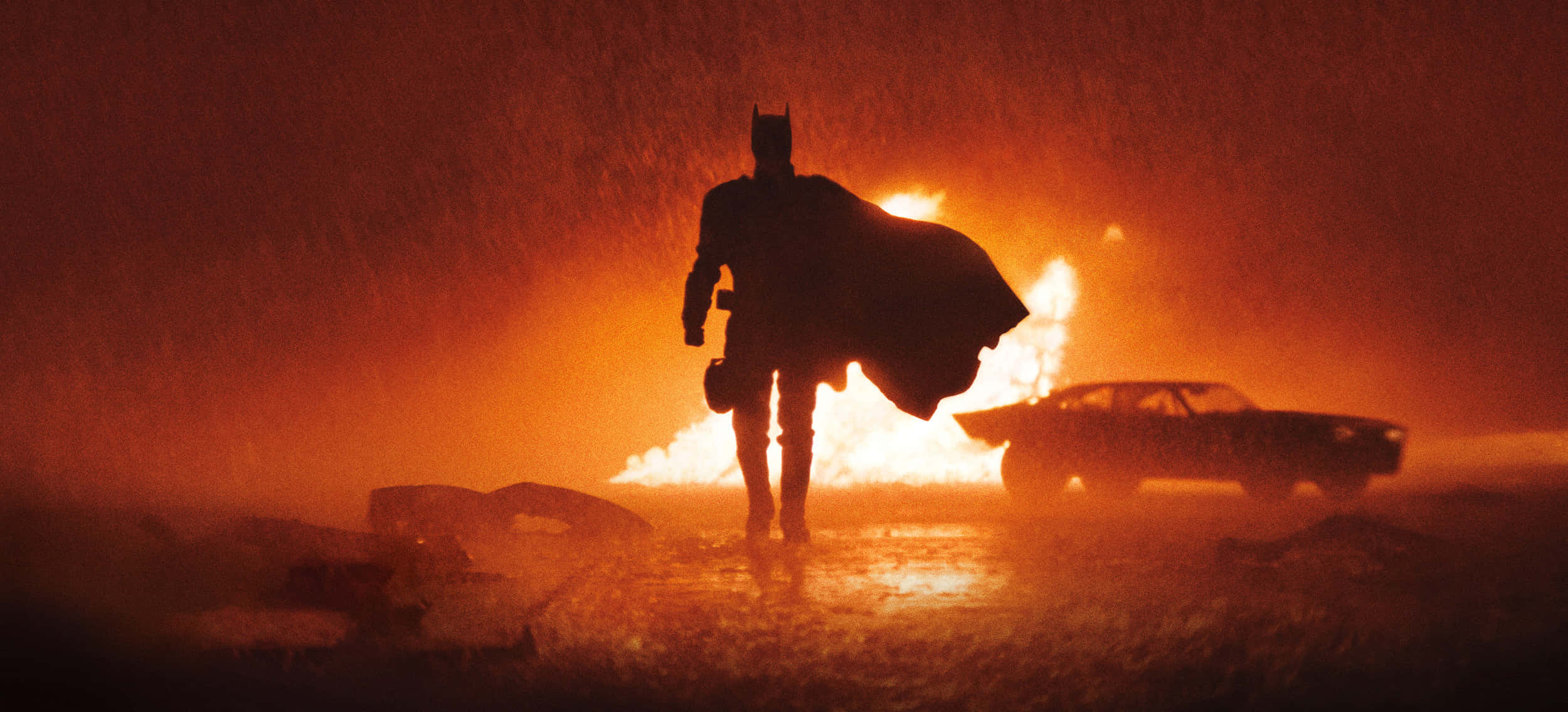 Batmobile Desktop 桌布 壁紙 — 蝙蝠俠在雨中背對著熾熱的橙色火焰和燃燒的汽車，形成剪影。