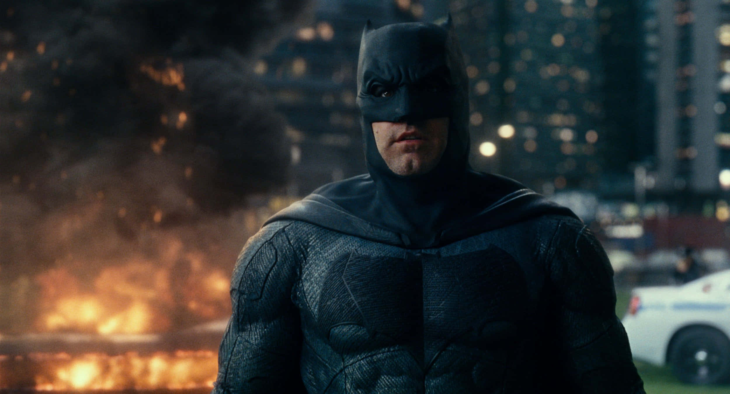Bat-suit 桌布 壁紙 — 蝙蝠俠在黑暗的城市景觀中堅定站立，夜燈下火焰和煙霧在他身後升起。