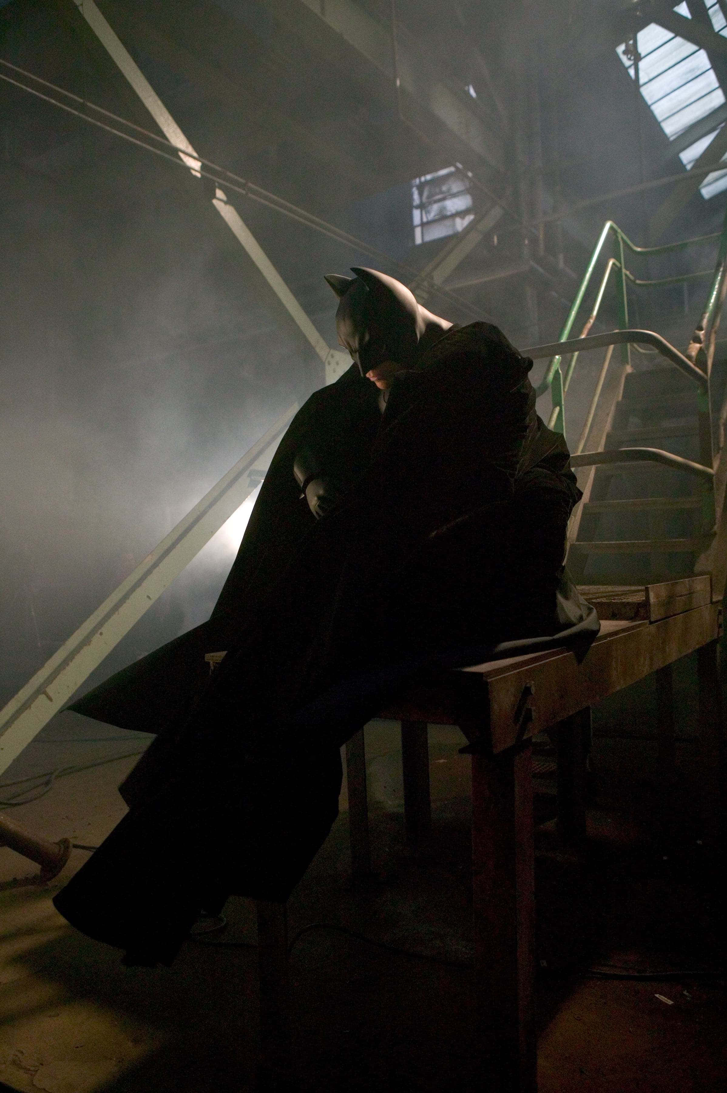 Batman Begins 桌布 壁紙 — 蝙蝠俠獨自坐在昏暗的工業空間中，沐浴在戲劇性的背光和煙霧中，散發出靜默的強烈感。