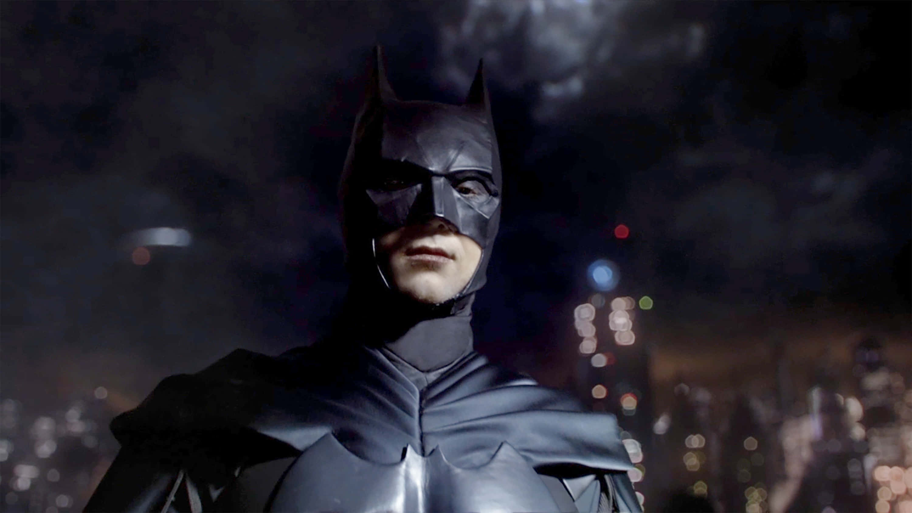 Bat-suit 桌布 壁紙 — 蝙蝠俠站在夜晚的黑暗城市景觀中，身後有模糊的燈光。