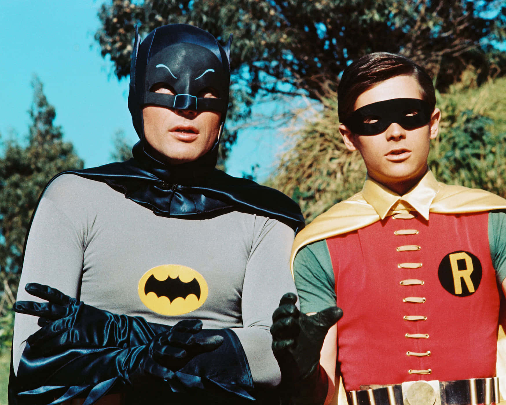 Batman And Robin 桌布 壁紙 — 蝙蝠俠與羅賓身穿經典1960年代服裝並肩站立，背後是明亮的藍天和綠樹。
