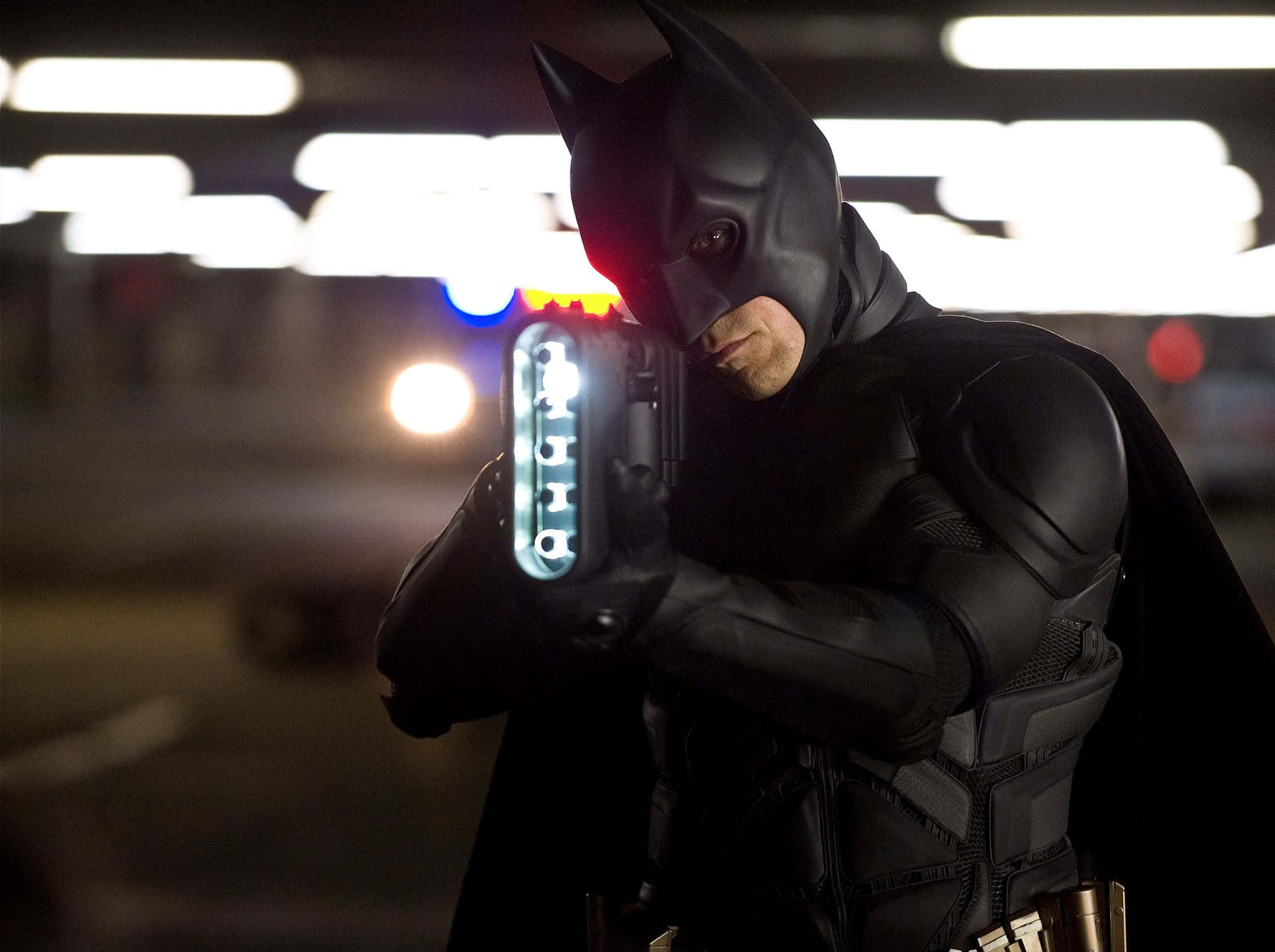 Bat-suit 桌布 壁紙 — 蝙蝠俠身穿暗色頭罩和戰衣，在光線昏暗的停車場中瞄準發光的蝙蝠鏢，背後有模糊的警燈。