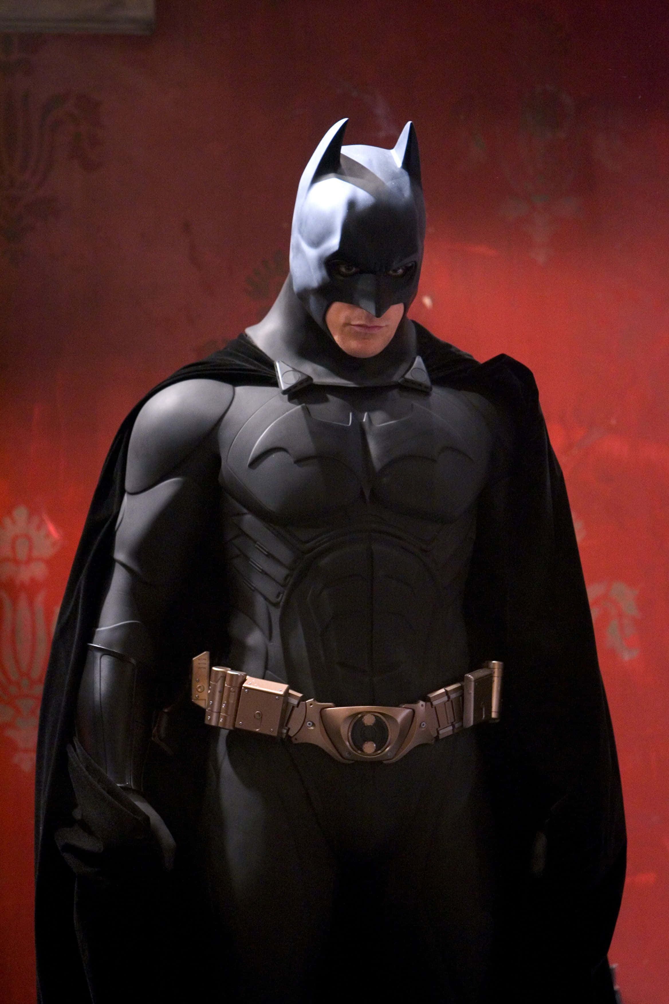 The Caped Crusader 桌布 壁紙 — 蝙蝠俠身穿深色西裝，站在帶有華麗圖案的紋理紅牆前，斗篷飄揚，眼神銳利。